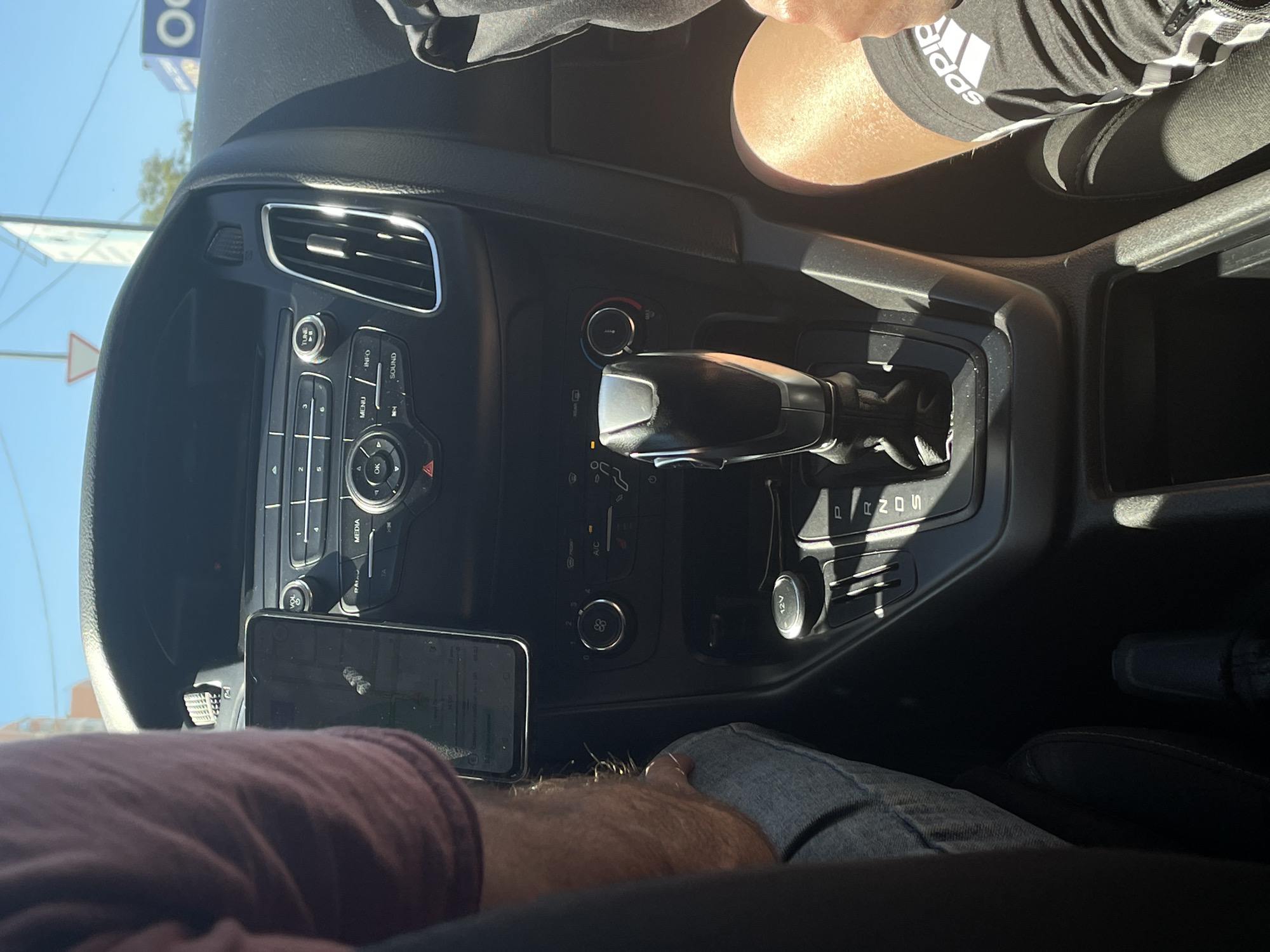 The image, taken inside a car in Rostov-on-Don, Russia, shows a view of the car's interior focusing on the central console and dashboard. Two individuals are partially visible. On the left, a person wearing a purple shirt and blue jeans is seated, with their arm resting on the console. A smartphone is mounted on the dashboard to their left, displaying a navigation app. On the right, another person is visible, wearing black Adidas shorts. The central console features an automatic gear shift, climate control buttons, and a 12V power outlet. The dashboard includes air vents and various buttons for the car's audio system. Outside the car, a clear sky and a building are visible through the windshield, suggesting a daytime scene on a sunny day.