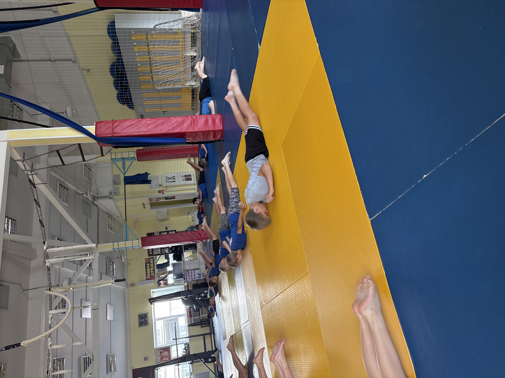 In a gymnasium in Chaikovsky, Russia, a group of young boys are lying prone on a blue and yellow mat. The boys, dressed in blue t-shirts and dark shorts, are arranged in rows, with their faces down and arms extended forward. The gymnasium features a high ceiling with exposed beams and industrial lighting. Ropes and climbing equipment are visible in the background, suggesting it is a facility for gymnastics or similar athletic training. The overall environment is that of an indoor sports facility.