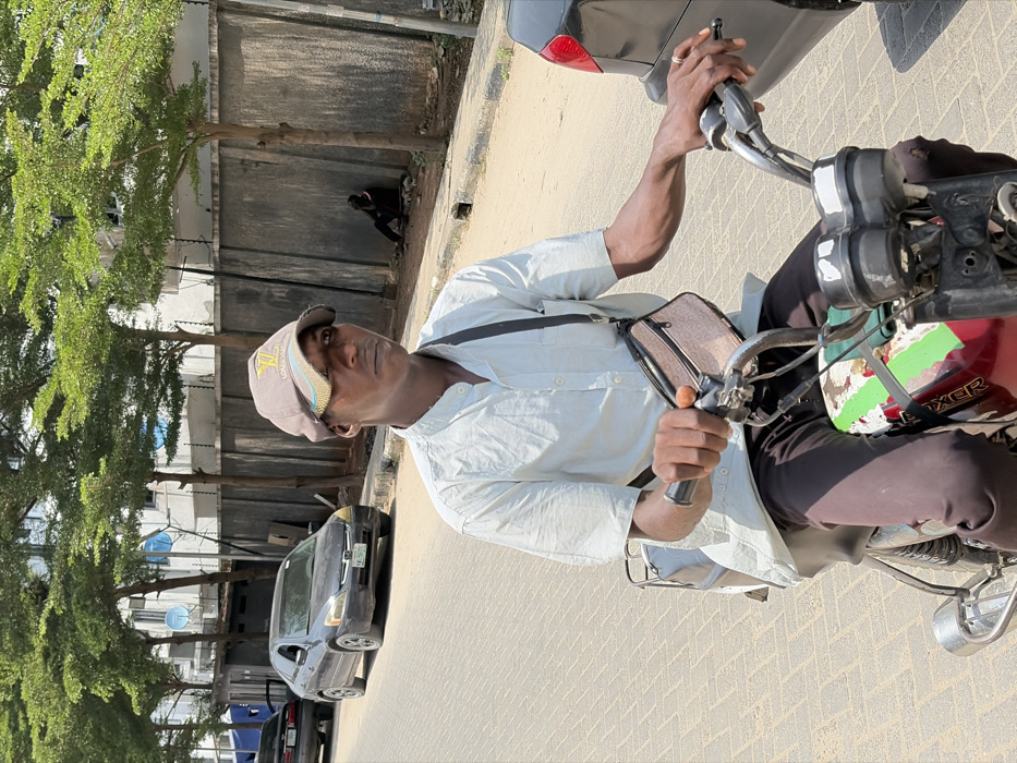A male individual is depicted in the foreground, seated on a red "BOXER" motorcycle on a paved street in Igboefon, Nigeria. He wears a light-colored, short-sleeved collared shirt, dark trousers, and a brown baseball cap with a gold-colored "LV" logo. A patterned fanny pack is slung across his chest. His hands grip the motorcycle's handlebars, with the right hand near the brake lever. The motorcycle's fuel tank shows areas of chipped paint and green/white markings. In the midground, to the left, a dark Honda Accord sedan with license plate "KM 565..." is visible on the street, followed by a partially visible dark vehicle. To the far right, the rear section of another dark vehicle is present. A concrete sidewalk with a drainage opening runs parallel to the street. In the background, a second individual, dressed in dark clothing, sits on the ground leaning against a tall, grey concrete wall topped with security wire. Multiple green-leafed trees line the road and the wall. White multi-story buildings with visible satellite dishes stand beyond the wall. The scene is illuminated by bright daylight, casting distinct shadows.