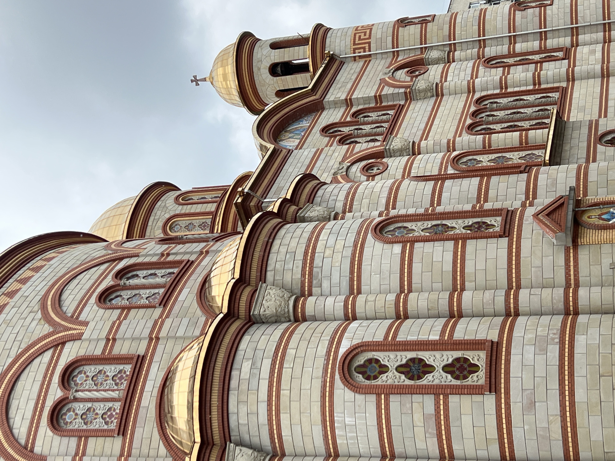 The image presents a detailed, low-angle view of a grand and ornate architectural structure, likely an Eastern Orthodox church, under an overcast sky. The building features multiple golden domes, some smooth and highly reflective, while others show tiled patterns. A prominent dome near the top left is surmounted by a small cross.
The facade is characterized by alternating bands of light-colored rectangular tiles and reddish-brown brickwork, often separated by thin, vertical golden strips. Numerous arched and rounded window-like recesses adorn the walls, many containing intricate stained-glass-like patterns in blues, reds, and yellows, set within the reddish-brown frames. Decorative elements, including carved stone details and a Greek key (meander) pattern, are visible on various sections of the building. The intricate layering and curving forms create a sense of depth and complexity.
The cloudy sky provides a soft, diffused light, accentuating the textures and colors of the building materials. No people or activities are visible, and the focus remains entirely on the architectural splendor. No discernible text is visible anywhere in the image.