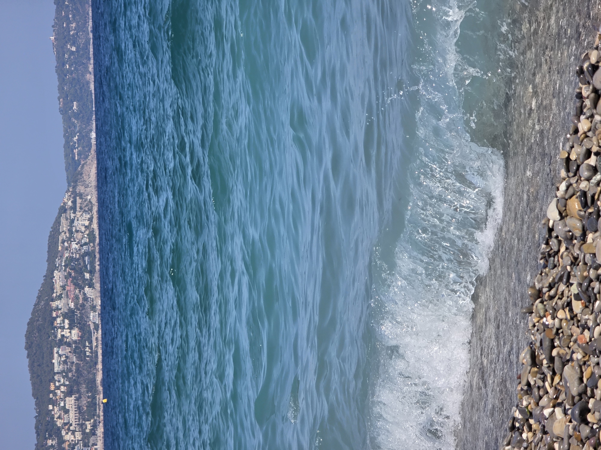 The image shows a view of the Mediterranean Sea from a pebble beach in Nice, France. In the foreground, a wave is breaking on the shore, with the clear blue water reflecting the sunlight. The beach is covered in smooth, colorful pebbles. In the background, a hillside is covered with buildings and vegetation, characteristic of the city of Nice. The sky is a clear, bright blue.