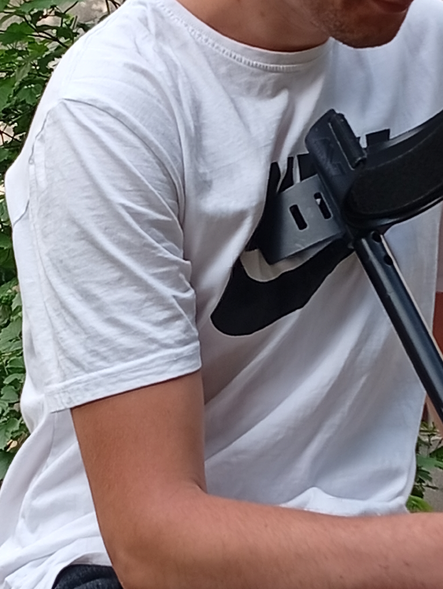 A close-up, partial image depicts the upper body of a person, likely male, positioned outdoors against a backdrop of green foliage. The individual wears a plain white short-sleeved t-shirt, which bears a prominent black logo resembling the Nike "swoosh" on the chest. Part of a dark-colored object, possibly a crutch or a similar support device, is visible resting against the person's right shoulder and chest area, extending downwards towards the lower right. This object features several rectangular cutouts in its structure. The person's bare right forearm is visible in the lower right portion of the frame. The background consists of vibrant green leaves and branches, suggesting a natural outdoor setting during daytime. The person's face is not visible, and their specific activity cannot be determined beyond being present. The scene is located in Poltava, Ukraine.