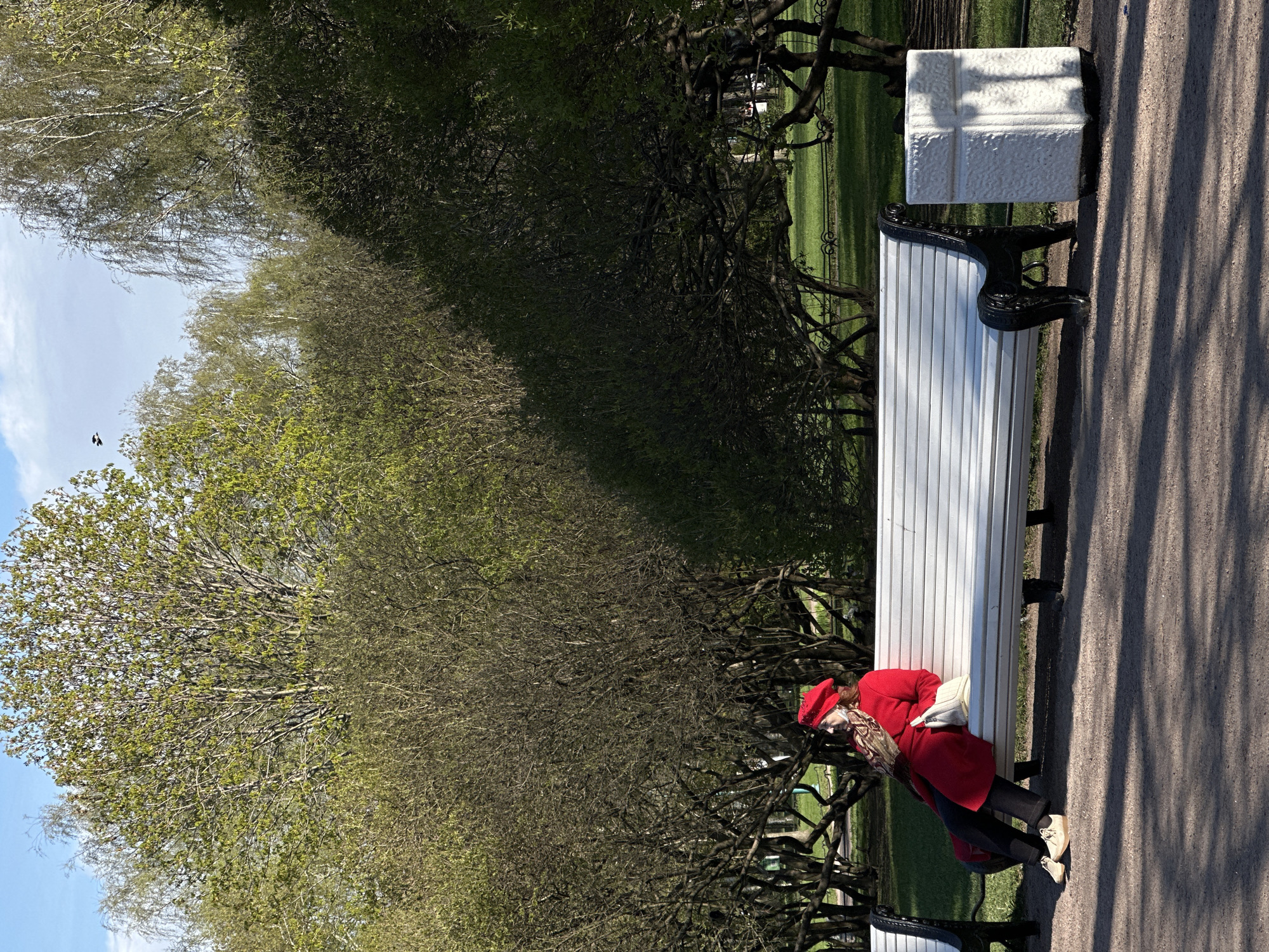 The image shows a park scene with a woman resting on a white park bench. She is wearing a bright red coat and matching hat. Her head rests on a white purse or bag, and she is wearing dark trousers and light-colored shoes. The bench is located on a pathway covered in gravel or small stones, casting long shadows. Beside the bench is a decorative white stone pillar. Behind the bench are numerous trees with new spring leaves, with a patch of green grass visible further back. The sky is blue with some clouds.