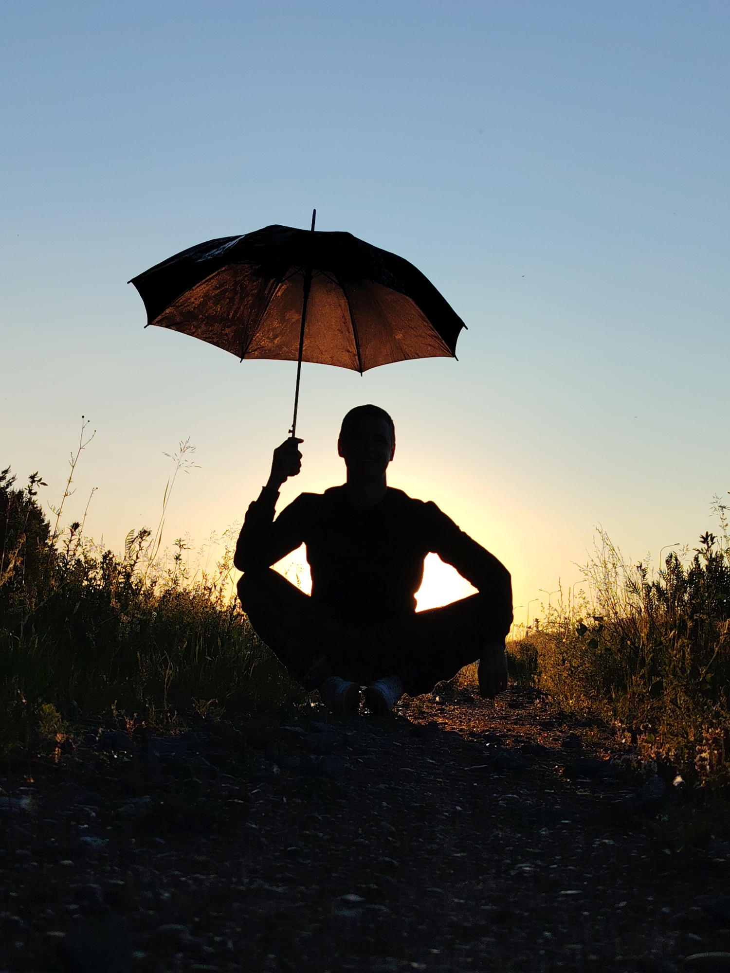 A silhouette of a person is crouched on a gravel path, holding an open black umbrella above their head. The sun is setting behind the person, casting a warm glow through the translucent umbrella and silhouetting the figure. The background consists of tall grass and vegetation on either side of the path, with a clear sky transitioning from pale blue at the top to a warm yellow-orange near the horizon. The scene appears to be outdoors, possibly in a park or a rural area, given the natural vegetation and lack of urban structures, and is a typical sunset view found in locations such as Chaikovsky, Russia.