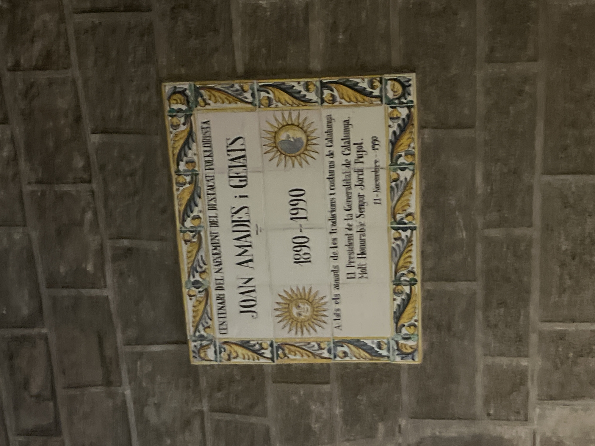 The image shows a decorative tile plaque embedded in a brick wall. The plaque is composed of multiple smaller tiles. The main inscription is in Catalan and commemorates the centenary of the birth of Joan Amades i Gelats, a prominent folklorist (1890-1990). It also mentions a dedication to those who love the traditions and customs of Catalonia, and specifically honors Jordi Pujol, the President of the Generalitat (regional government) of Catalonia at the time, with the date November 11, 1990. The plaque features decorative elements, including stylized suns. The surrounding wall is made of dark gray or brown bricks. The location is very likely Barcelona, Spain, given the Catalan language and the mention of the Generalitat of Catalonia.
