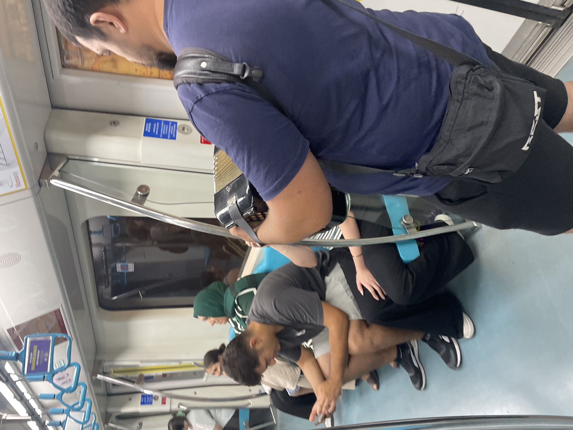 The image depicts the interior of a metro train in Istanbul, Turkey. Several passengers are seated on blue seats. In the foreground, a man wearing a navy blue t-shirt and black shorts is playing an accordion. He has a black crossbody bag slung over his shoulder. To his left, a young man in a dark gray t-shirt and light gray shorts is looking down at his phone. Further down the row, a woman wearing a green hijab is seated. Other passengers are visible in the background. The train carriage has white walls and ceiling, with blue grab handles for standing passengers. The floor is a light blue color.