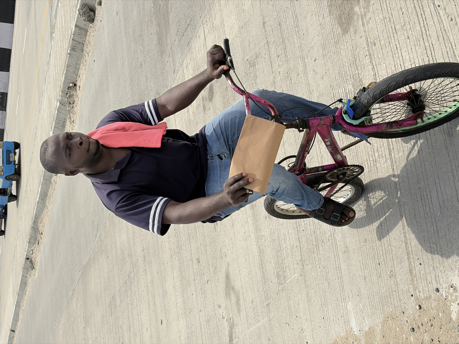 A male subject, estimated to be in his 20s to 30s, with short dark hair and dark skin, is depicted riding a bicycle on a concrete road in Jakande, Nigeria. He is wearing a dark blue short-sleeved collared shirt with two white horizontal stripes on each sleeve, light blue denim jeans with visible wear, and dark open-toed sandals. A red towel is draped over his left shoulder and neck. He holds a rectangular, light brown, flat object in his left hand; a silver ring is visible on his left pinky finger. His right hand grips the right handlebar of the bicycle. The subject's gaze is directed towards the upper right of the frame.
The bicycle is a small, BMX-style model, predominantly painted a faded and chipped magenta/pink. Its wheel rims are a distinct light green or aqua color, and a light blue plastic piece is attached to the front fork area.
The environment consists of a wide, light-colored concrete road. To the left side of the frame, in the background, a raised concrete curb separates the current road section from another paved area where two light blue, three-wheeled utility vehicles are visible in the distance, traveling away from the viewer. Scattered small debris is present on the road surface. Strong shadows are cast on the ground, indicating direct overhead sunlight.