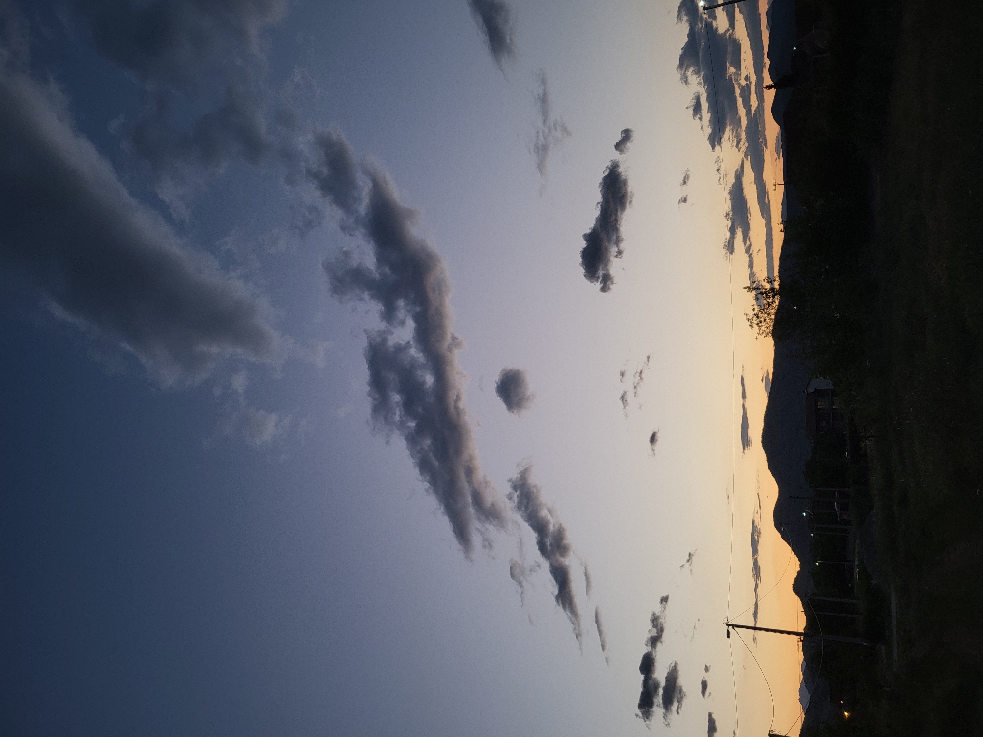 This photograph depicts a serene natural landscape at either dusk or dawn, characterized by a striking gradient in the sky. The upper left portion of the sky is a deep, dark blue, transitioning smoothly towards warmer orange and yellow hues at the bottom-right, indicative of the setting or rising sun. Scattered across this vast sky are numerous dark, elongated clouds, some appearing wispy and others more dense, silhouetted against the lighter parts of the sky.

Along the horizon, primarily in the lower half of the image, a chain of mountains or hills is visible in dark silhouette against the vibrant sky. The foreground and midground areas are mostly dark, showing indistinct land, possibly fields or grassy areas, with trees and bushes also appearing as dark outlines. Several utility poles with power lines stretch horizontally across the lower portion of the frame, and a few artificial lights are faintly glowing, reinforcing the impression of evening twilight. Distant buildings are also subtly visible on the hillsides, adding a touch of human presence to the natural setting. The scene evokes a sense of tranquility and the calm transition from day to night, in a rural or semi-rural area like Mahala, Montenegro. No people or animals are visible, and there is no discernible text.