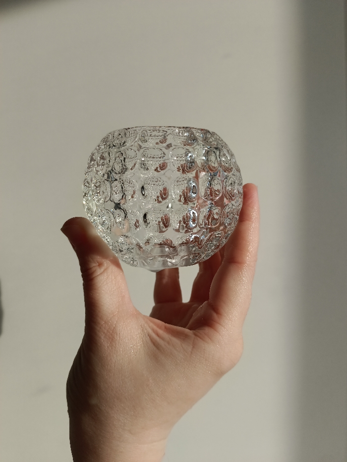 An adult human hand with light skin tone is depicted holding a clear glass object against a plain, light-colored background. The hand is positioned centrally, holding the object from below and partially from the sides with the thumb and index finger visibly supporting it. The glass object is spherical in shape, featuring a uniform textured pattern of convex, circular dimples or bubbles covering its exterior. It appears to have an open top, suggesting it may be a votive holder or small vase. Strong directional lighting from the left illuminates the scene, highlighting reflections and refractions within the glass and creating subtle shadows on the right side of the background. The background itself is featureless, appearing to be an indoor wall. The specific location within Chaikovsky, Russia, is not discernible from the visual evidence presented in the image.