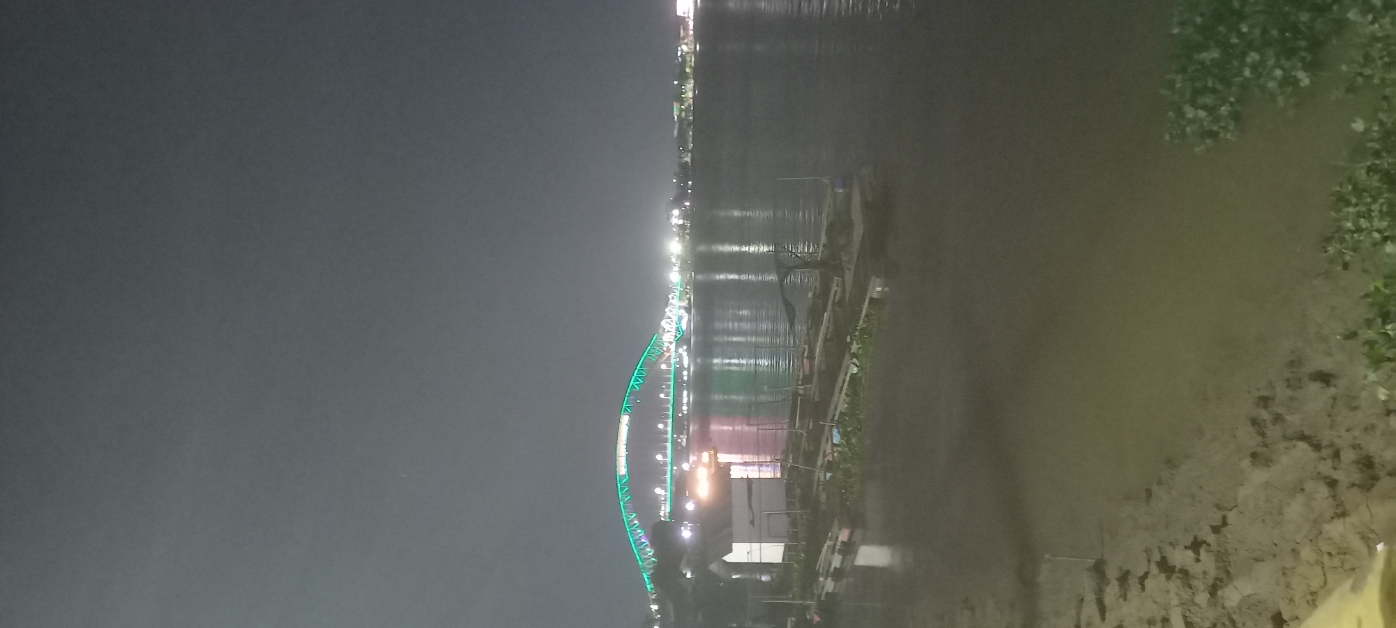The image shows a nighttime view of the Teluk Dalam bridge in Teluk Dalam, Indonesia, illuminated by green lights. The bridge spans a body of water, likely the sea, and is reflected on the surface. On the left side of the foreground, there is a small white building on stilts over the water, and several small boats or rafts are moored nearby. The shoreline is visible in the lower left of the image, with some vegetation. The background shows a city skyline with various lights. The overall atmosphere is dark and somewhat hazy, typical of a nighttime scene.