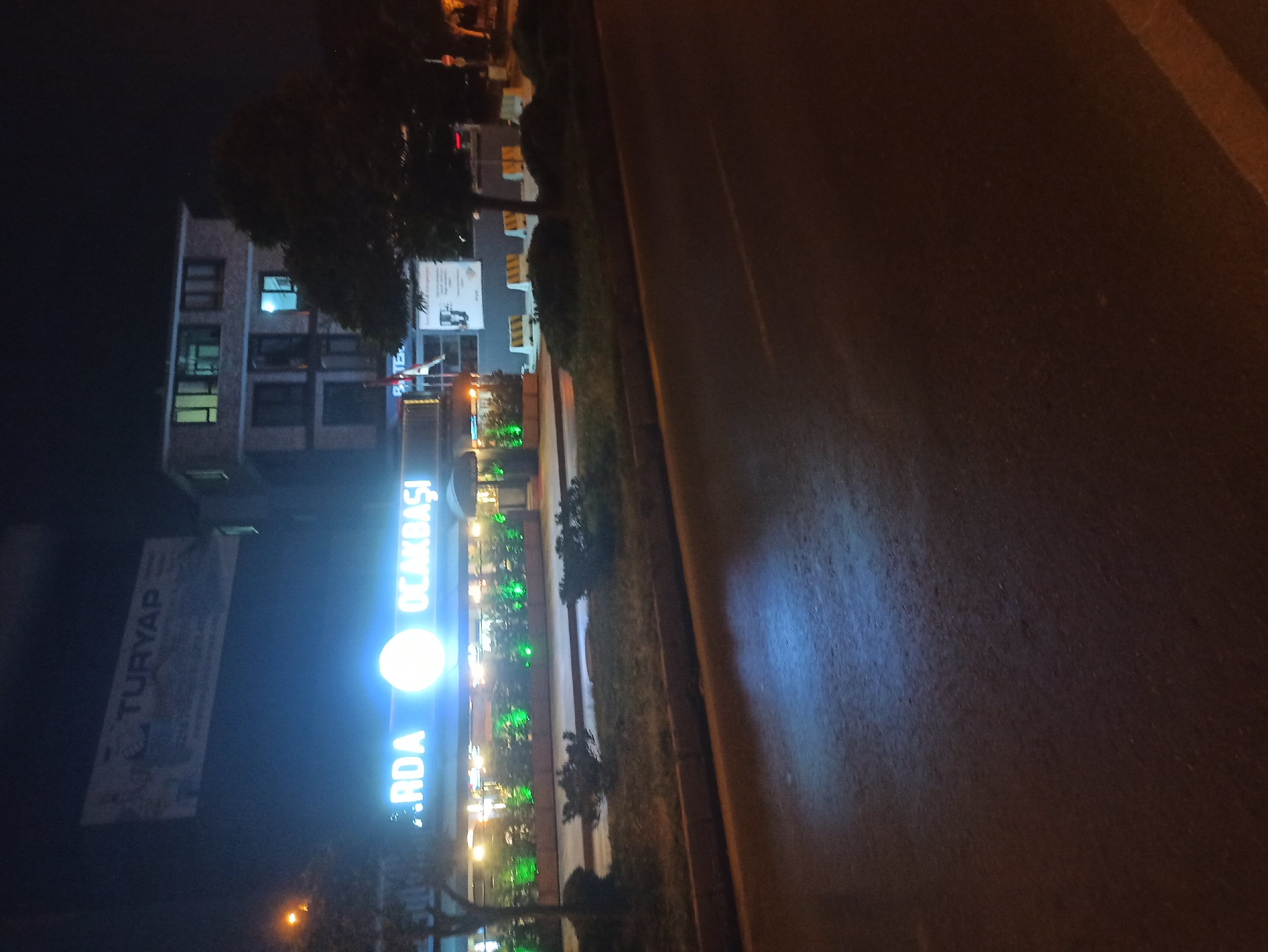The image captures a nighttime street scene in Ümraniye, Turkey, specifically featuring the entrance of a restaurant or business named "ARDA OCAKBAŞI." The establishment is illuminated by bright, neon-blue signage and green outdoor lighting highlighting greenery along its facade. Above the "ARDA OCAKBAŞI" sign, a larger, rectangular billboard is visible, displaying "TURYAP" in prominent lettering, suggesting a real estate or property agency. Behind the restaurant, a multi-story building with illuminated windows is present. The street in front of the establishment appears wet, reflecting the lights from the building and the road. The foreground is dominated by a dark asphalt road with a faint white line marking. Trees and bushes are present along the sidewalk, with some construction barriers or yellow and white plastic dividers visible further down the street. The overall atmosphere is that of a commercial area at night.