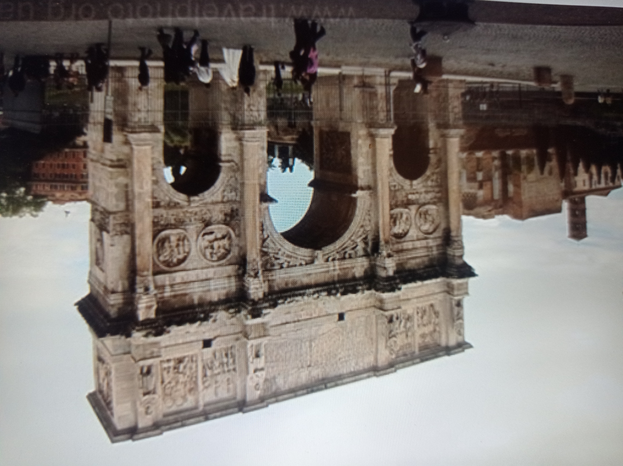The image shows the Arch of Constantine in Rome, Italy, not Mukachevo, Ukraine. It is a grand, ancient triumphal arch made of stone, adorned with intricate carvings and sculptures depicting historical scenes. The arch has a large central archway flanked by smaller archways. In front of the arch, there is a paved area with several people walking around. The sky is overcast. In the background, there are other ancient Roman ruins and buildings, including what appears to be a tower on the left. The overall impression is one of historical significance and grandeur.