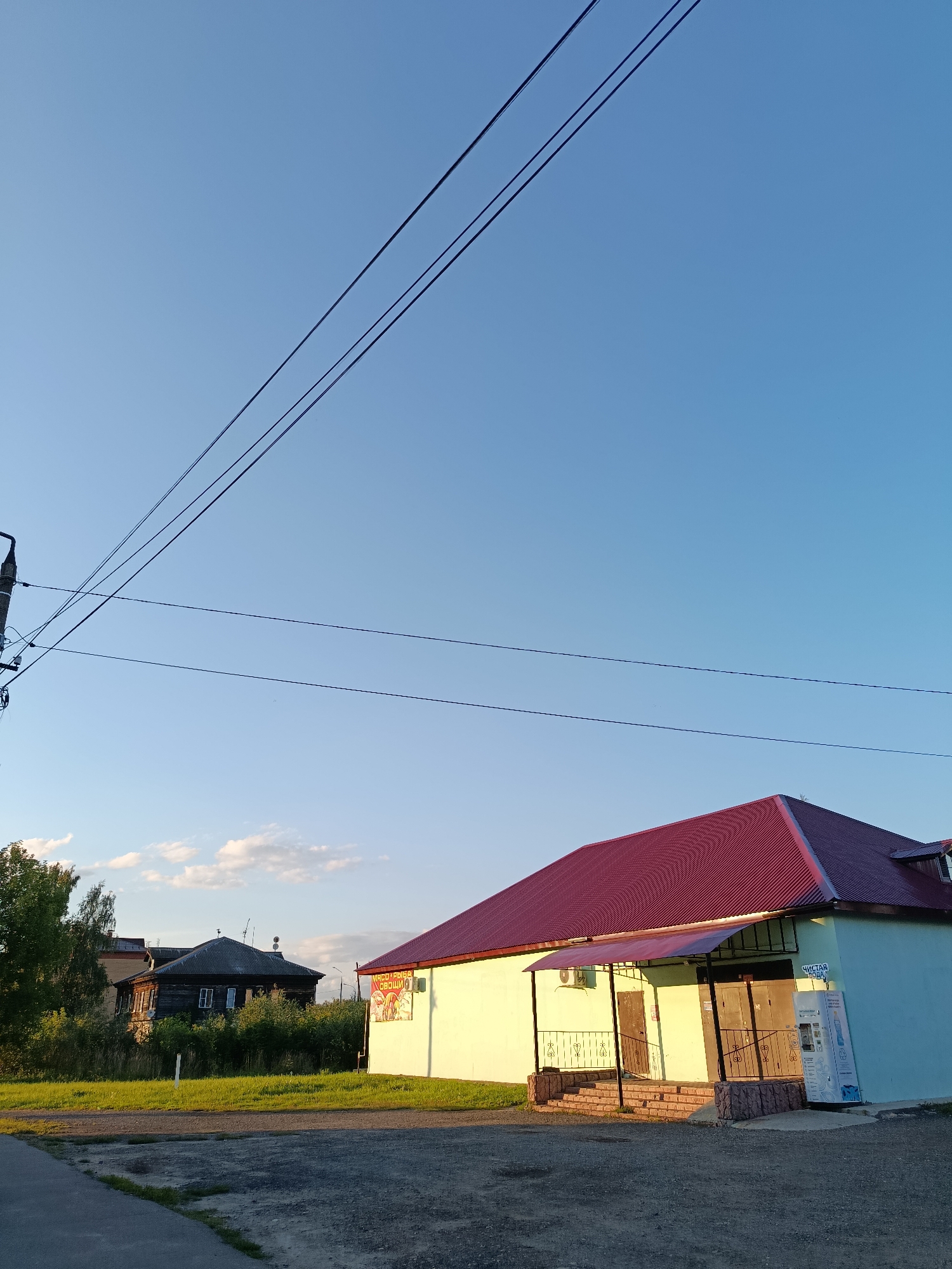 The image shows a street scene in the city of Vereya, Russia, under a clear blue sky. In the foreground, a light-colored building with a burgundy roof is visible. The building has an awning over its entrance, which features a set of stairs and a railing. To the right of the entrance, a vending machine for bottled water is positioned against the building's wall. Further in the background, a dark, weathered wooden house with white-trimmed windows is surrounded by dense green foliage. Electrical wires are strung across the upper portion of the image, supported by a pole visible on the far left. The ground in front of the buildings is a gravel and asphalt surface, with a paved walkway on the left. A sign with Cyrillic lettering is attached to the side of the light-colored building. The scene appears to be illuminated by the soft light of either early morning or late afternoon sun. There are no people or animals visible in the image.