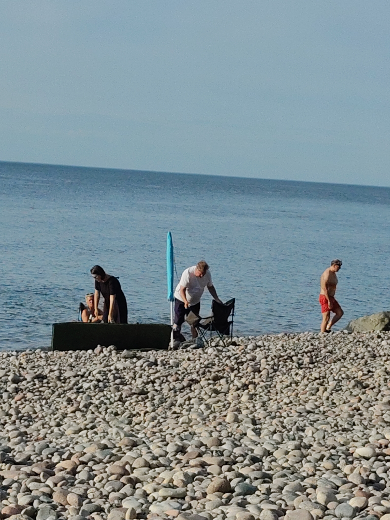This image depicts a scene on a pebbly beach next to the sea, likely in Pionersky, Russia, given the context. In the foreground, a vast expanse of grey and tan pebbles covers the beach. The sea, a calm blue, stretches to the horizon under a clear, pale blue sky.
Several people are present. On the left, a woman in a black dress is bending over, attending to someone who appears to be seated in a green container-like object near the water's edge. To their right, a man in a white t-shirt and dark pants is standing next to a folded black camping chair, looking down. Further to the right, a man wearing red swim trunks walks along the beach, away from the water and towards a large grey rock on the right side of the frame. A bright blue beach umbrella is partially visible standing upright between the group on the left and the man with the chair. The overall atmosphere is relaxed, suggesting a leisurely day at the beach.