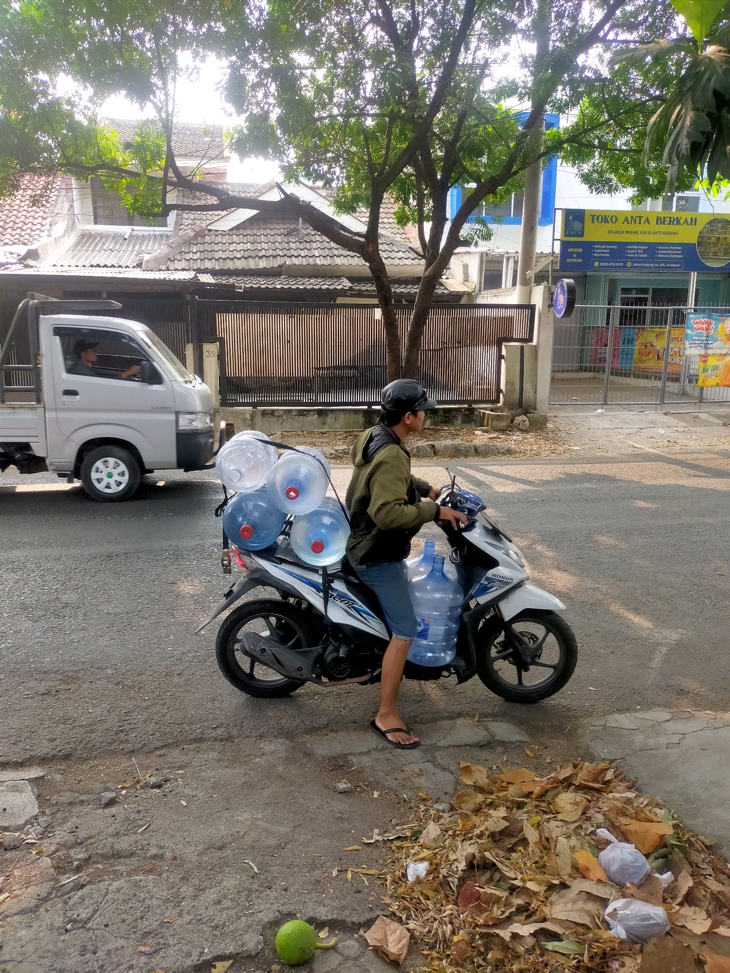 A person rides a white Honda motorcycle, carrying five large blue and clear water jugs on the back. The person wears a dark helmet, a green jacket, and blue shorts. They are on a paved street in front of a building with a blue and yellow sign that reads "TOKO ANTA BERKAH" and has text in Indonesian. To the left of the motorcycle, a silver truck with "CARRY" written on its side is visible, with a person driving it. The environment appears to be a street in a residential or commercial area, with trees and buildings in the background. A pile of dry leaves and some plastic bags are on the ground to the right of the motorcycle. A green fruit is on the ground in the lower left corner of the image. The scene suggests a delivery or transportation activity in a city.