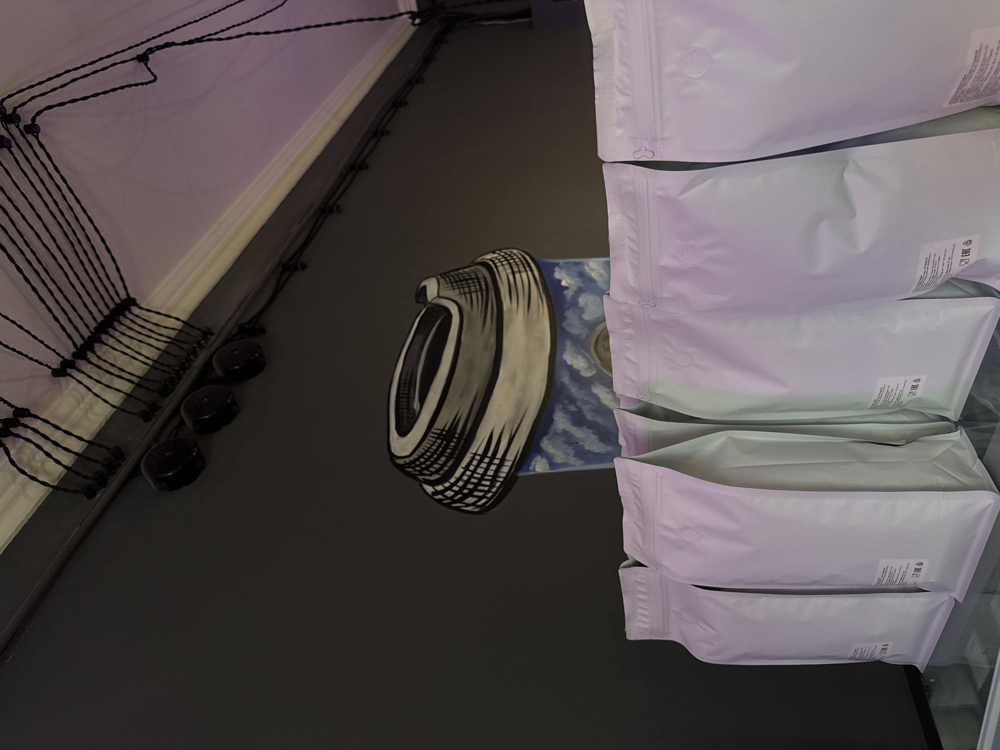 The image captures a close-up, angled view. The environment appears to be an interior space, possibly a shop or a storage area.
On the right side of the frame, there is a stack of white, sealed bags. The bags seem to be stacked vertically, partially obscuring the background. There are markings or labels on the bags, though the details are not fully discernible.
Behind the bags, there is a wall or surface with a decorative element. This includes a black and white stylized image and a blue and white section, possibly a painting or mural.
To the left, there are what appear to be electrical wires or cords, possibly part of a lighting setup. Further to the left, are round black objects that might be buttons.
Given the objects and the close-up nature of the image, it's difficult to pinpoint the exact location within Moscow, Russia without more context. The scene appears to be an interior of a business, possibly one that sells or stores packaged goods.