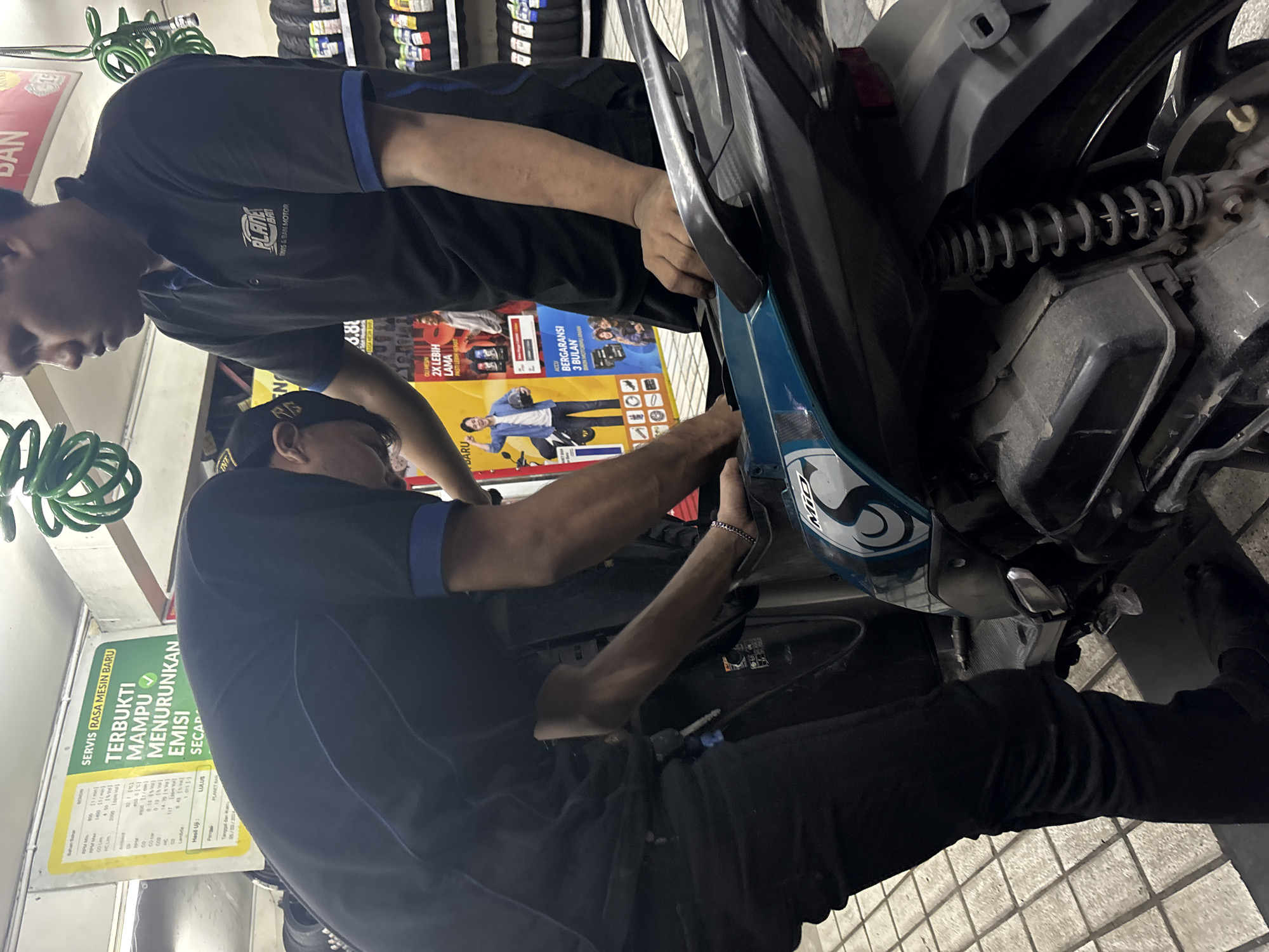 Two men are working on a blue and black scooter inside what appears to be a service center or shop. One man, wearing a black polo shirt with a blue collar and trim, and the "PLANET BAN" logo on the left chest, stands on the right, looking down at the scooter and has his right hand on the scooter's body. The other man, wearing a black t-shirt and black pants, is bent over the scooter, with his hands on the engine area. The scooter has a "Mio" decal on its side.
In the background, there is a green coil of hose hanging from the ceiling, a yellow and green sign with Indonesian text that reads "SERVIS RASA MESIN BARU" and "TERBUKTI MAMPU MENURUNKAN EMISI SECARA," and a shelf with stacked tires. The floor is tiled with square gray tiles. The overall scene suggests a motorcycle maintenance or repair service. The "PLANET BAN" branding suggests the location is a Planet Ban outlet, a known tire and automotive service chain in Indonesia, with a significant presence in cities like North Jakarta.