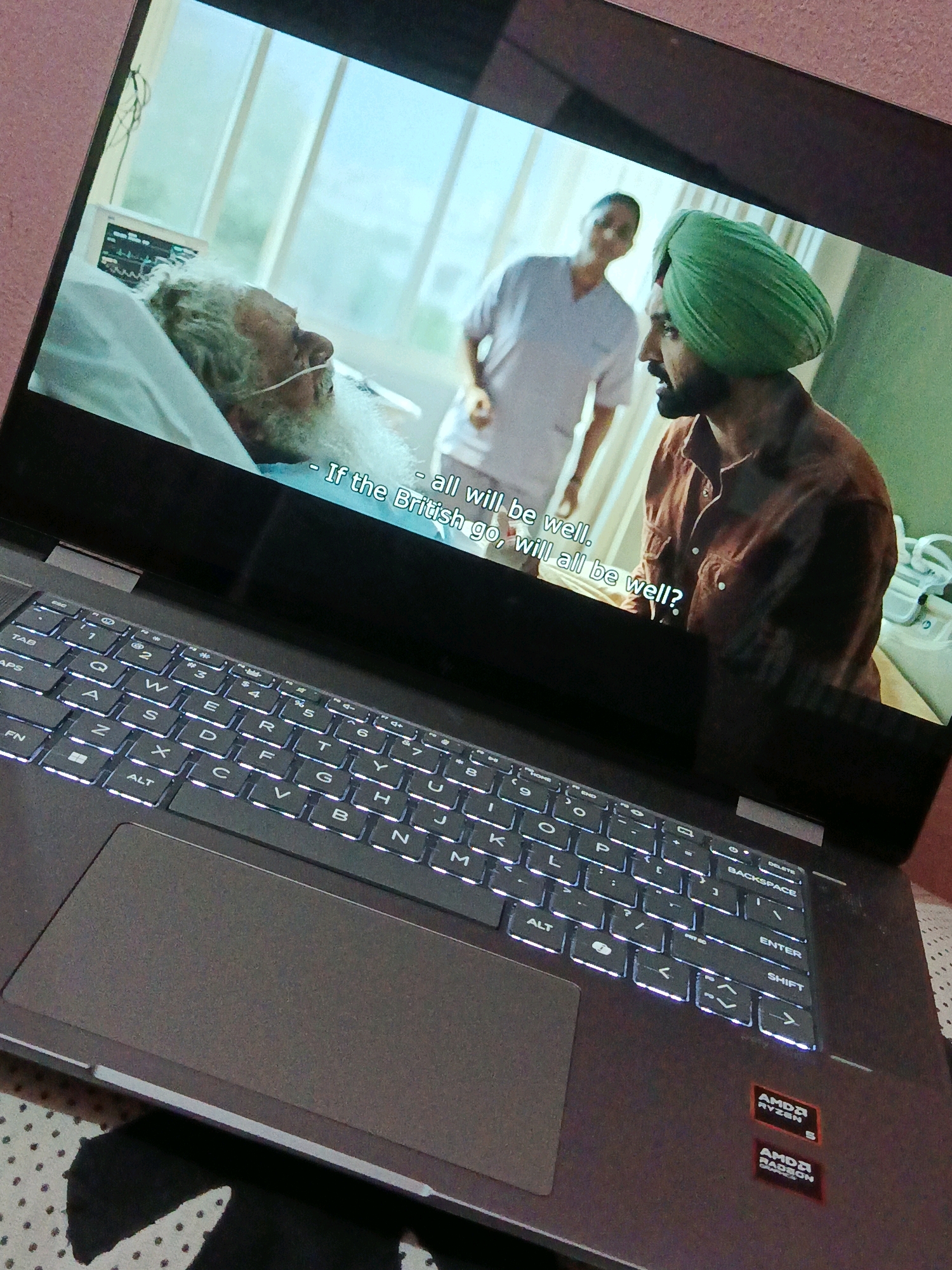 A laptop is open, displaying a scene from a movie or television show. The screen shows three people in what appears to be a hospital room. In the foreground, an elderly man lies in a hospital bed, connected to medical monitoring equipment. He has a long white beard and is wearing an oxygen tube. Behind him, a person in a light-colored uniform stands, and to the right, a man wearing a green turban and a brown shirt is visible in profile, speaking. Subtitles on the screen read, "- If the British go, will all be well?". The laptop keyboard is illuminated, with keys such as TAB, CAPS, FN, ALT, Q, W, E, R, T, Y, U, I, O, P, A, S, D, F, G, H, J, K, L, Z, X, C, V, B, N, M, SHIFT, ENTER, and BACKSPACE clearly visible. The overall setting suggests an indoor environment. There is no indication of the city of Dhaka, Bangladesh, in the image.
