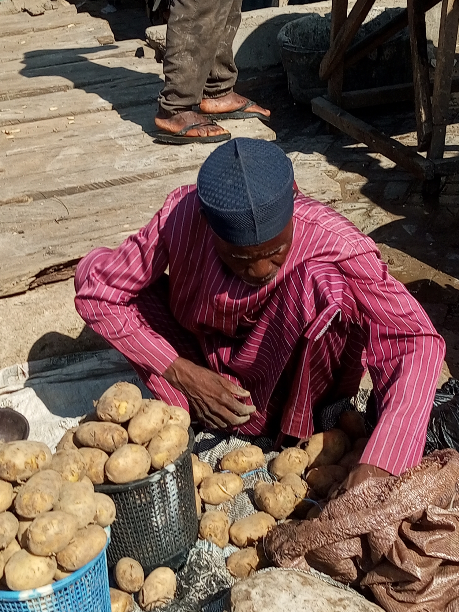 An overhead shot captures a Nigerian man, dressed in a magenta and white striped robe and a dark blue cap, crouching amidst a market stall. His hands are clasped together, partially obscured by the fabric of his robe, as he appears to be tending to a pile of potatoes. The potatoes are mostly light brown, some with visible blemishes, and are arranged in woven baskets and directly on the ground. Behind him, another individual's legs are visible, clad in muddy brown trousers and sandals, standing on a weathered wooden platform. The environment suggests an outdoor marketplace, possibly in Tempure, Nigeria, given the wooden structures and earthy tones indicative of a rural or semi-urban setting. The lighting indicates a bright, sunny day, casting distinct shadows. The scene is raw and unvarnished, focusing on the mundane act of commerce and everyday life.