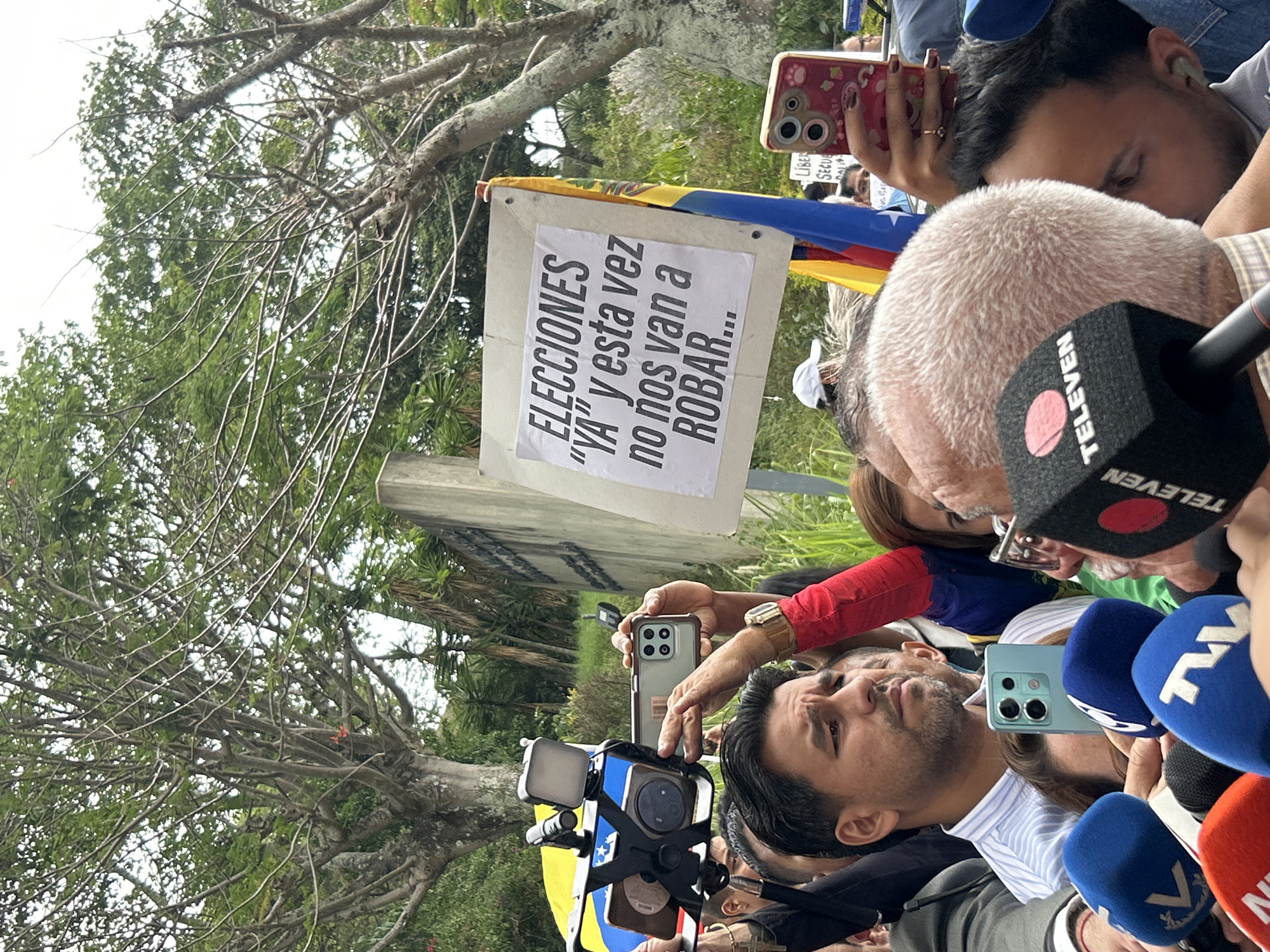 This image captures an outdoor political gathering, likely a protest or press conference, in Caracas, Venezuela. The scene shows several individuals actively participating, with a strong focus on media coverage.
In the foreground, multiple people are gathered closely. A man with dark hair and a beard is prominent, looking slightly upwards, surrounded by several microphones from different media outlets, including one clearly marked "TELEVEN." This suggests he is either speaking to the press or is a central figure at the event. Other participants are seen holding up smartphones, actively recording the proceedings, indicating widespread engagement and documentation of the event.
A significant detail is a white sign, held aloft, with bold black text in Spanish. It reads: "ELECCIONES 'YA' y esta vez no nos van a ROBAR." This translates to "Elections 'NOW' and this time they will not ROB us," conveying a strong political message demanding immediate elections and expressing distrust regarding the electoral process. Partially visible behind the sign and among the crowd is a flag featuring yellow, blue, and red stripes, consistent with the Venezuelan flag, further anchoring the geographical and political context.
The setting is outdoors, characterized by bright daylight and green trees with some bare branches in the background, suggesting a public park or street location. The presence of numerous microphones and recording devices highlights the media attention surrounding this gathering, indicating a newsworthy event with strong public sentiment.