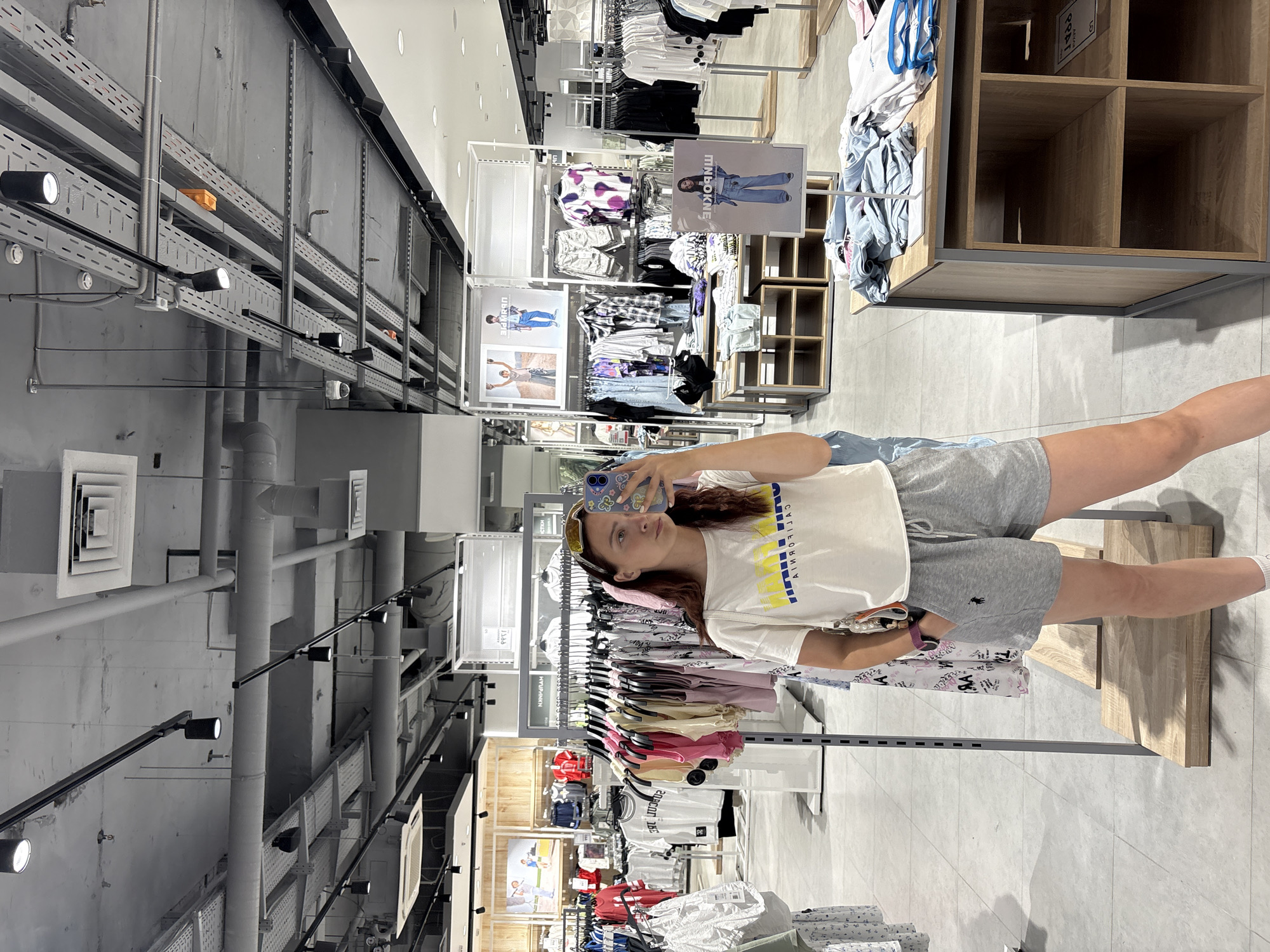 A young woman with long, dark hair tied back and wearing sunglasses on her head stands in a clothing store and takes a mirror selfie with her phone. She is wearing a white t-shirt with yellow and blue lettering, grey sweat shorts with a small black logo, and white socks. In the background, racks of colorful clothing are visible, along with store displays, shelving units, and a tiled floor. The ceiling features exposed pipes, ductwork, and lighting fixtures. The overall impression is a modern retail environment. The image appears to have been taken in Izhevsk, Russia, as indicated by the text on the t-shirt which reads "CALIFORNIA" in English, but the sign behind her has text in Cyrillic script which could indicate the location.