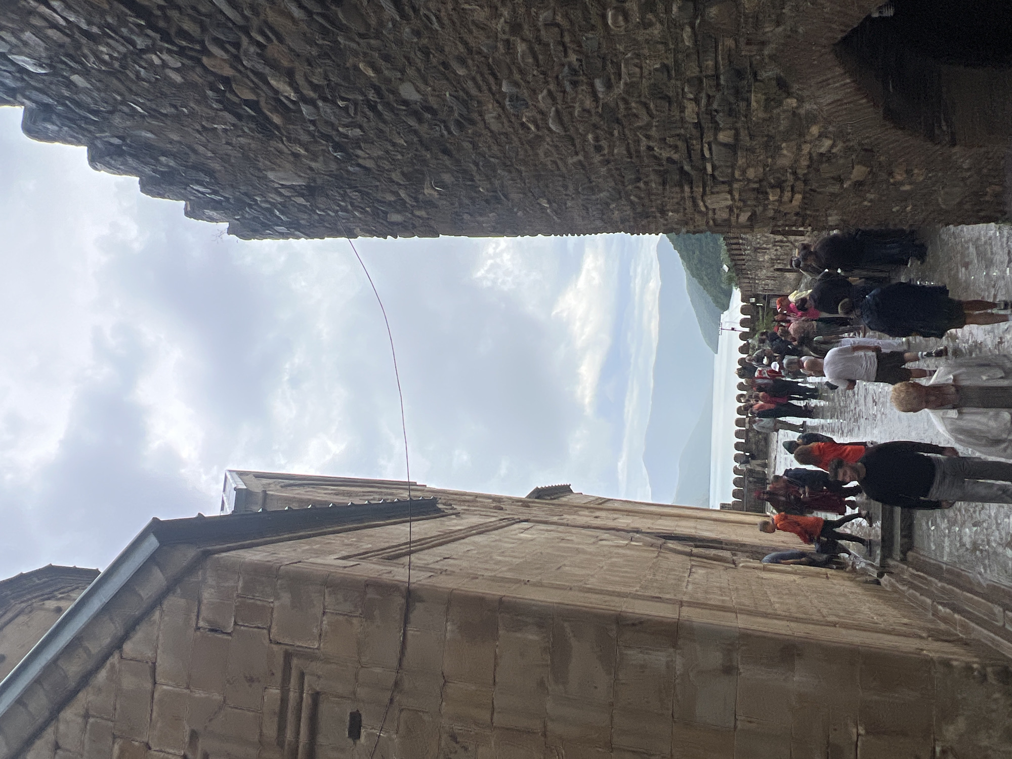 The image shows a group of tourists walking through a narrow passage between two ancient stone structures, possibly part of a historical complex in Shalikiantkari, Georgia. The passage opens up to a scenic view of a large body of water, possibly a reservoir or a lake, with distant mountains and a cloudy sky overhead. The ground is wet, suggesting recent rain. The people are dressed in casual clothing, and some are carrying umbrellas, indicating a possibly overcast or rainy day. The architecture of the buildings appears to be old, constructed from large stone blocks. The overall atmosphere is that of a tourist destination with a historical and natural appeal.
