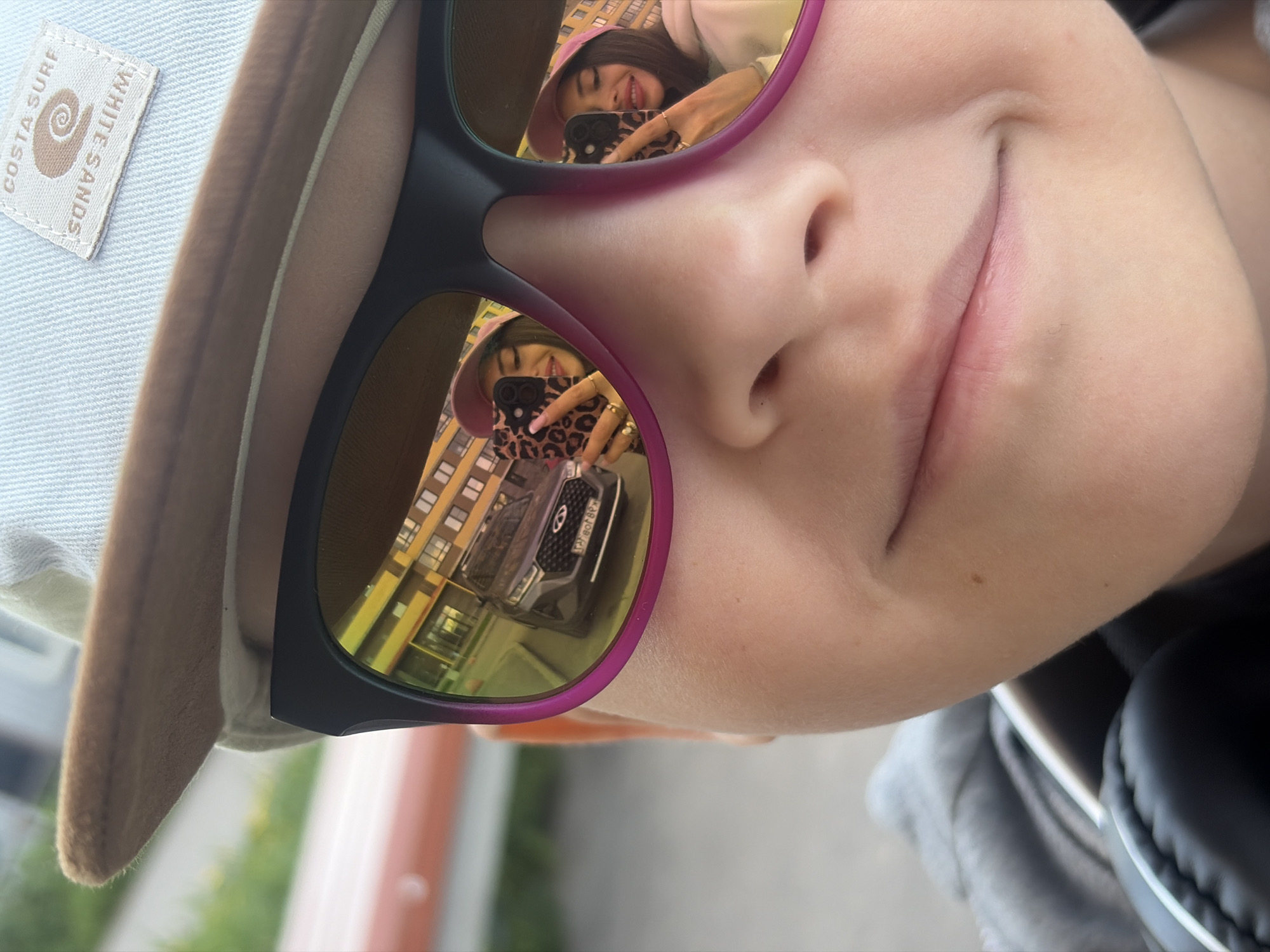 The image is a close-up, slightly tilted portrait capturing the face of a young person wearing a light blue or off-white bucket hat with a brown brim and mirrored sunglasses. The person's face is positioned on the left side of the frame, showing part of their nose and a gentle, upward-curving smile. They appear content.
The most prominent features are the mirrored lenses of the sunglasses, which reflect a scene. In the left lens, a woman is clearly reflected, wearing a pink bucket hat and holding a smartphone with a distinctive leopard print case. She is smiling and appears to be taking a photo or selfie, with multiple rings visible on her fingers. Behind her, a multi-story urban building with numerous windows is reflected, along with a dark grey SUV (likely a Hyundai) parked, bearing a Russian license plate. The right lens reflects a similar, but less distinct, image of the woman, the building, and the car.
The hat features a fabric label with the text "COSTA SURF" and "WHITE SANDS" around a spiral or shell logo. The reflected car's license plate, when de-mirrored, reads "AO 338 E 178," with "178" being the region code for Saint Petersburg, Russia, confirming the provided location context.
The background behind the person in the foreground is softly blurred, showing hints of green foliage and muted building tones, suggesting an outdoor setting during daylight hours. The person also wears black over-ear headphones, visible on the right side of the frame, and a grey hooded garment. The overall mood is casual and pleasant, likely a moment captured during an outing.