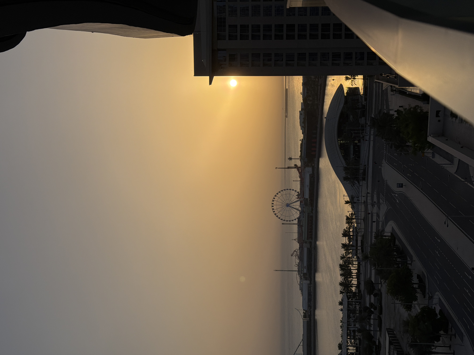 The image depicts a sunset view from a high vantage point in Lusail, Qatar. The scene is dominated by the warm hues of the setting sun, casting a golden glow across the sky and water. In the foreground, a modern building with many windows is partially visible on the right, silhouetted against the bright sky. A balcony railing runs across the bottom of the frame, reflecting the sunlight.
Further into the distance, across a body of water, an amusement park with a prominent Ferris wheel and roller coaster tracks is visible. The waterfront is lined with palm trees and several buildings, including a distinctive modern structure with a curved roof. A large ship can be seen sailing on the water.
The lower part of the image shows a cityscape with roads, streetlights, and more palm trees. The overall impression is one of a modern, developing city at dusk, with the natural beauty of the sunset enhancing the urban landscape. There are no people visible in the image.