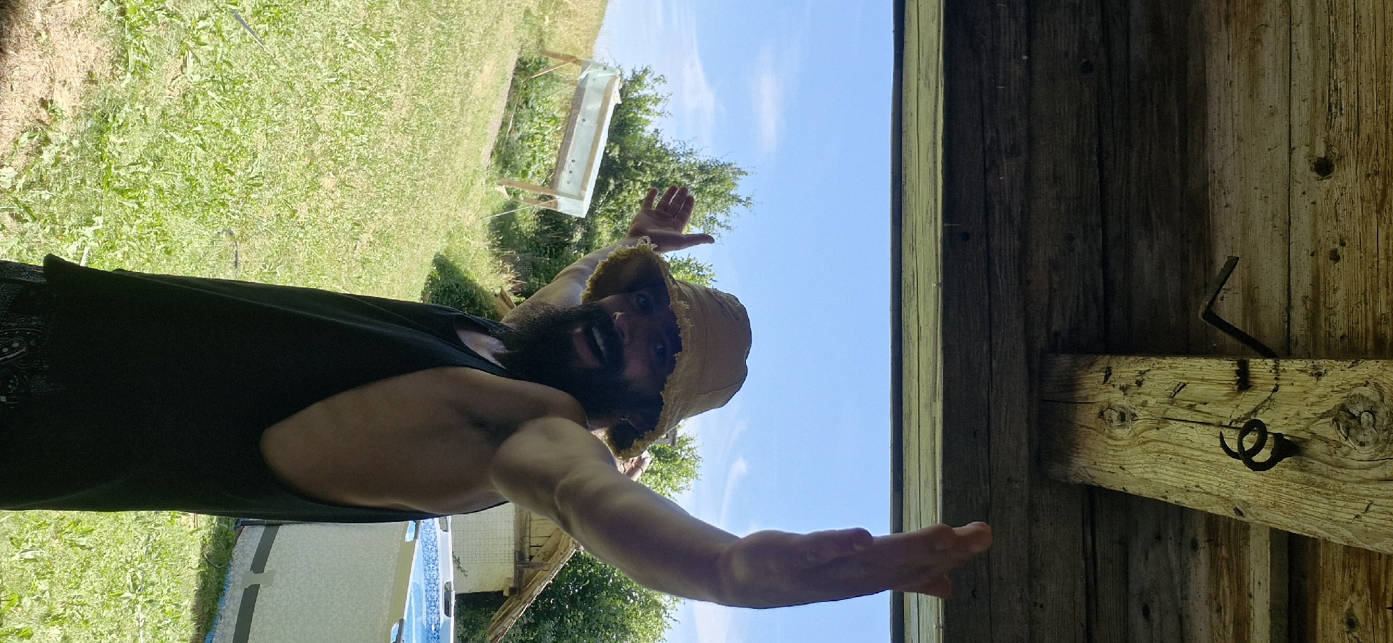 A man with a beard and a bucket hat is looking at the camera with a wide smile, holding out his hands. He is wearing a black tank top. The man is outdoors, with a grassy yard and a blue sky visible. There is a wooden structure to the right of the image, possibly a shed or porch. The scene appears to be set during the daytime, with bright sunlight. The man's expression conveys happiness and excitement. No text is visible in the image.