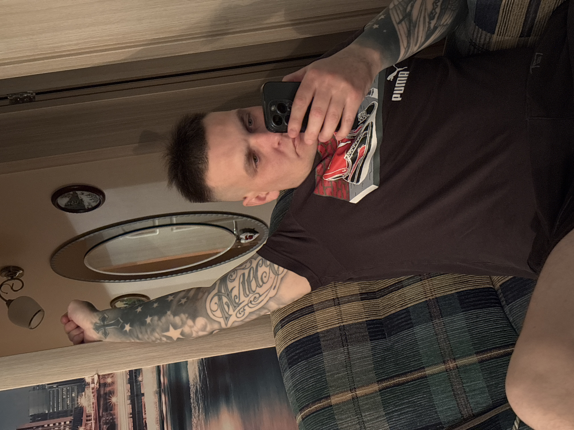 A man is indoors, in what appears to be a home setting, possibly in the city of Русский Вожой, Russia. He is holding a smartphone, presumably taking a selfie. The man is wearing a dark brown Puma T-shirt and dark shorts. He has multiple tattoos on his left arm. In the background, there is a wall with a mirror and a wall decoration. He is sitting on a sofa with a plaid pattern. The overall lighting is somewhat subdued.
