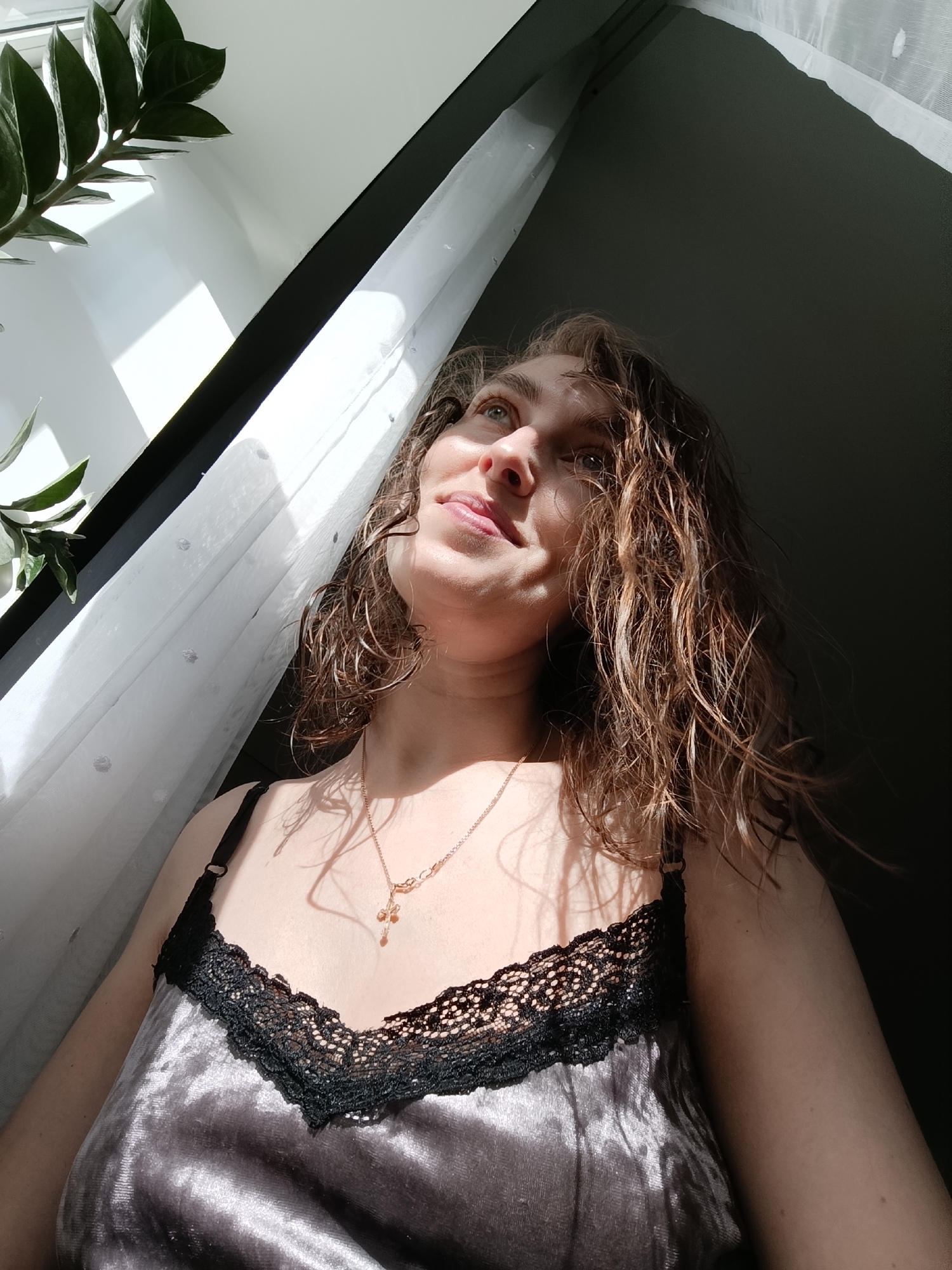 A woman with wavy, brown hair, is standing in front of a window with white, semi-transparent curtains. Sunlight streams in from the left side of the frame, partially illuminating her face and hair. She is wearing a gray camisole with black lace trim and a delicate gold necklace with a cross pendant. Her gaze is directed upward and slightly to the left, with a slight smile on her face. A green leafy plant is visible in the upper left corner of the frame. The background wall is a dark color, contrasting with the bright light from the window. This image does not contain enough information to discern the exact location in Chaikovsky, Russia.