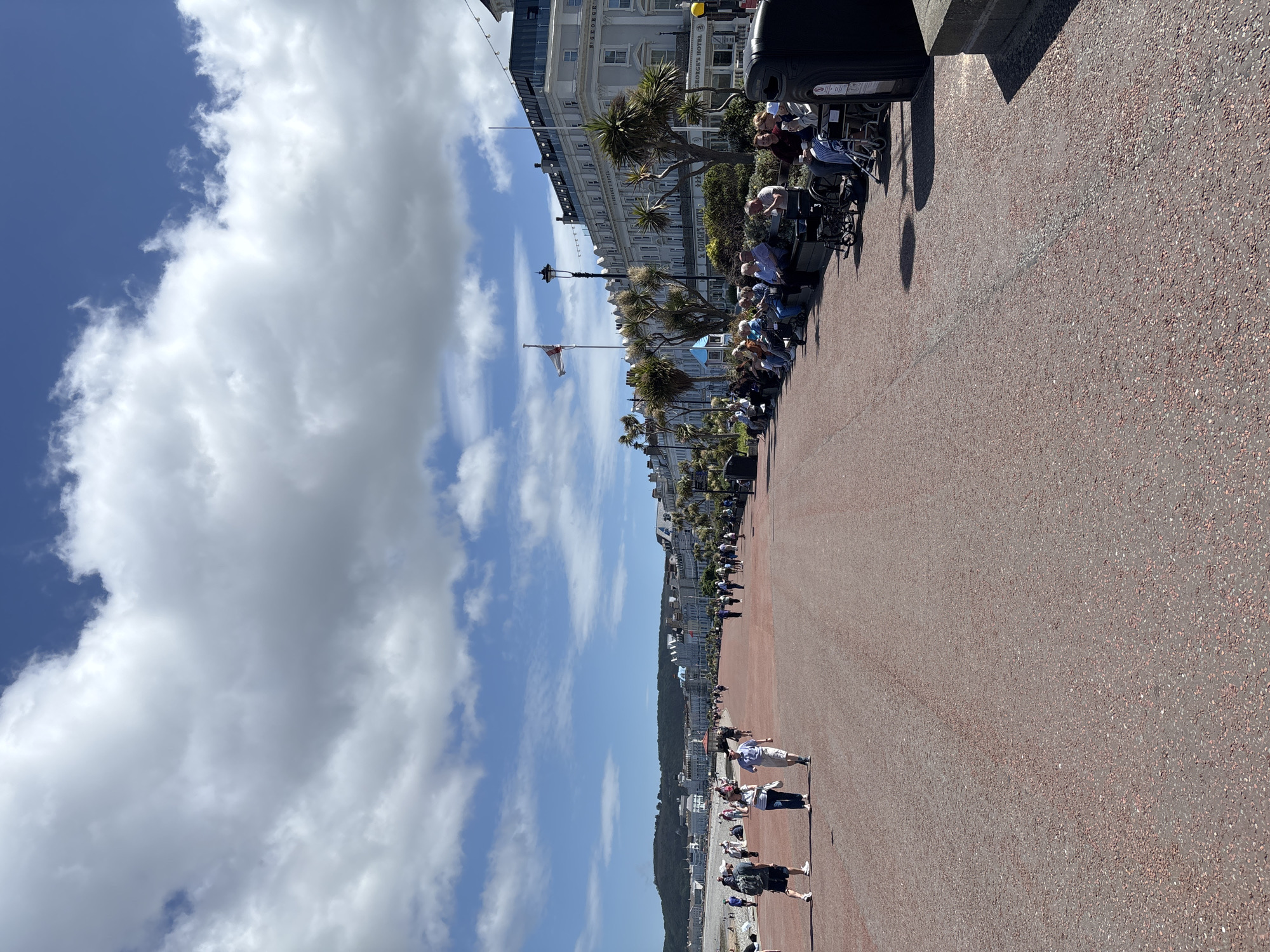 The image presents a view of the seafront promenade in Llandudno, United Kingdom, under a partly cloudy sky. A wide walkway, textured in a reddish-brown, dominates the foreground and extends into the distance. Along the edge of the walkway closest to the buildings, a series of benches are occupied by people seated and facing the sea. Some individuals appear to be in wheelchairs. The buildings lining the promenade are light-colored, multi-story structures featuring architectural details like balconies and awnings. Palm trees are interspersed along the building front. A flag is visible, flying from a pole in front of the buildings. In the distance, the coastline stretches with buildings and a backdrop of a wooded hill or small mountain. Some people can be seen walking and relaxing on the beach below the promenade.