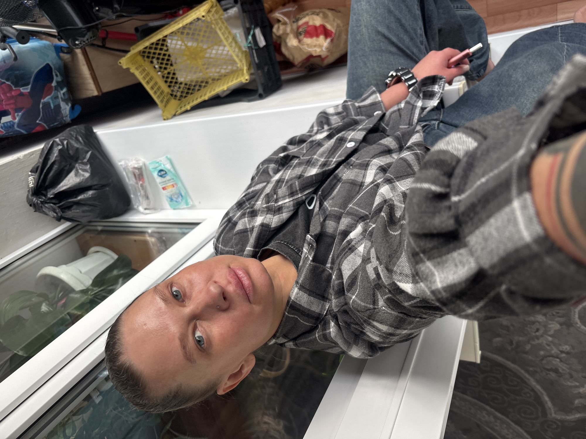 A person wearing jeans and a gray and white plaid shirt is holding a small pink device and wearing a watch. They are in Perm, Russia. A yellow plastic basket, black trash bag, cleaning supplies and plants are in the background. The person is sitting on or near a white surface.