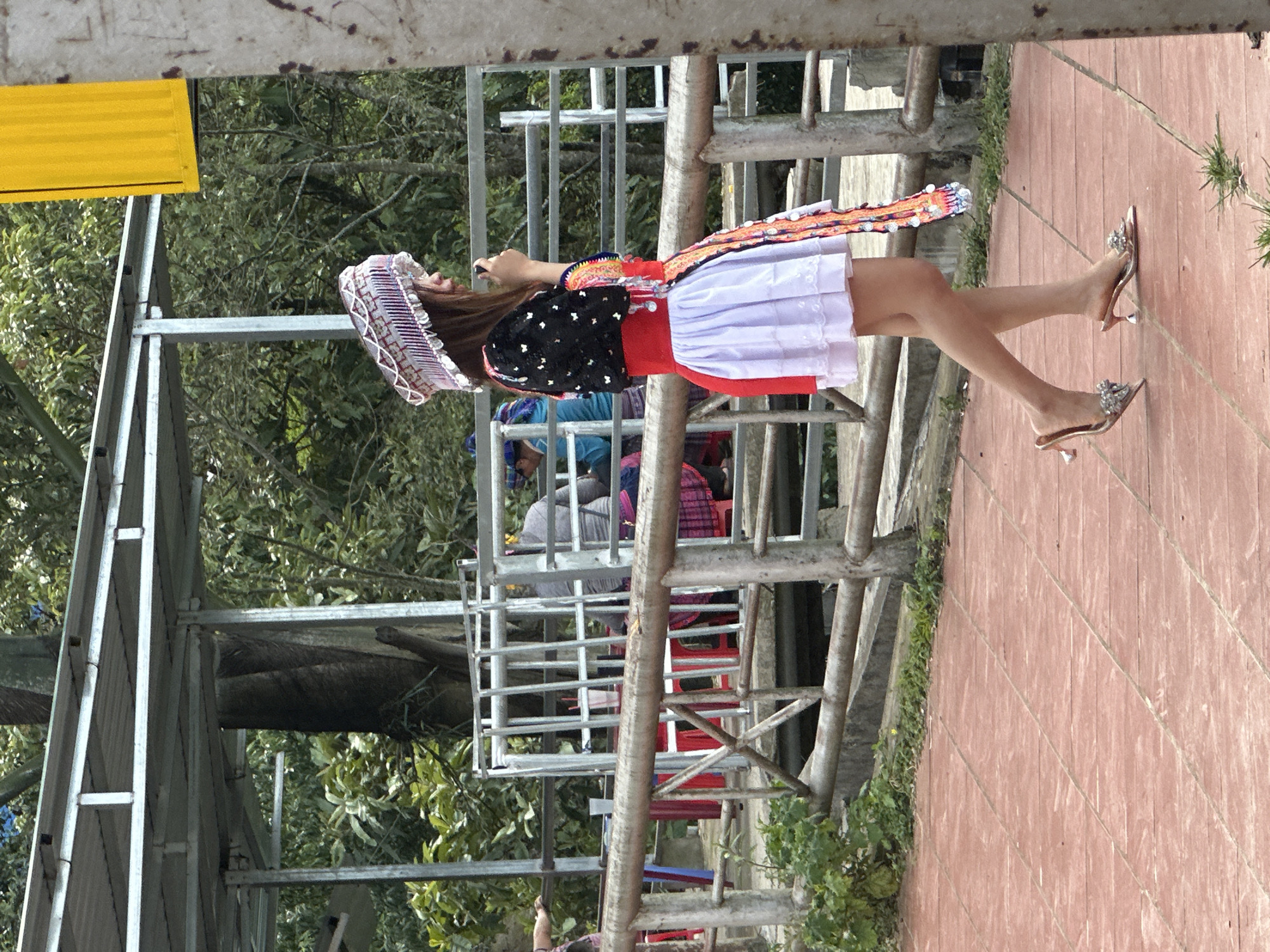In Tu Le, Vietnam, a young woman, facing left in profile, walks across a reddish-brown tiled pavement. She is dressed in traditional attire, featuring an elaborate patterned silver-and-white headpiece with dangling ornaments, a black short-sleeved top with white butterfly-like embellishments, a wide red waist belt, and a white pleated mini-skirt with a red panel on the left side. A long, colorful, beaded or sequined sash hangs from her left hip. She wears low, pointed-toe, high-heeled mules with sparkling embellishments on the toe strap. Her right hand holds a small dark object, possibly a phone or microphone, near her face.
Behind her, a concrete railing with metal posts and horizontal bars separates the walkway from an area below. Further back, an open-air structure with a corrugated metal roof supported by metal framework provides shelter. Beneath this structure, several individuals are visible, seated at tables with red plastic chairs. One person on the far left wears a blue top, and another to the right wears a grey top and a patterned pink skirt. Lush green trees and foliage dominate the background, with a large tree trunk visible centrally. On the far right, a partial view of a bright yellow rectangular sign or banner is visible next to a grey pillar. Green vegetation sprouts intermittently along the edge of the pavement and railing.