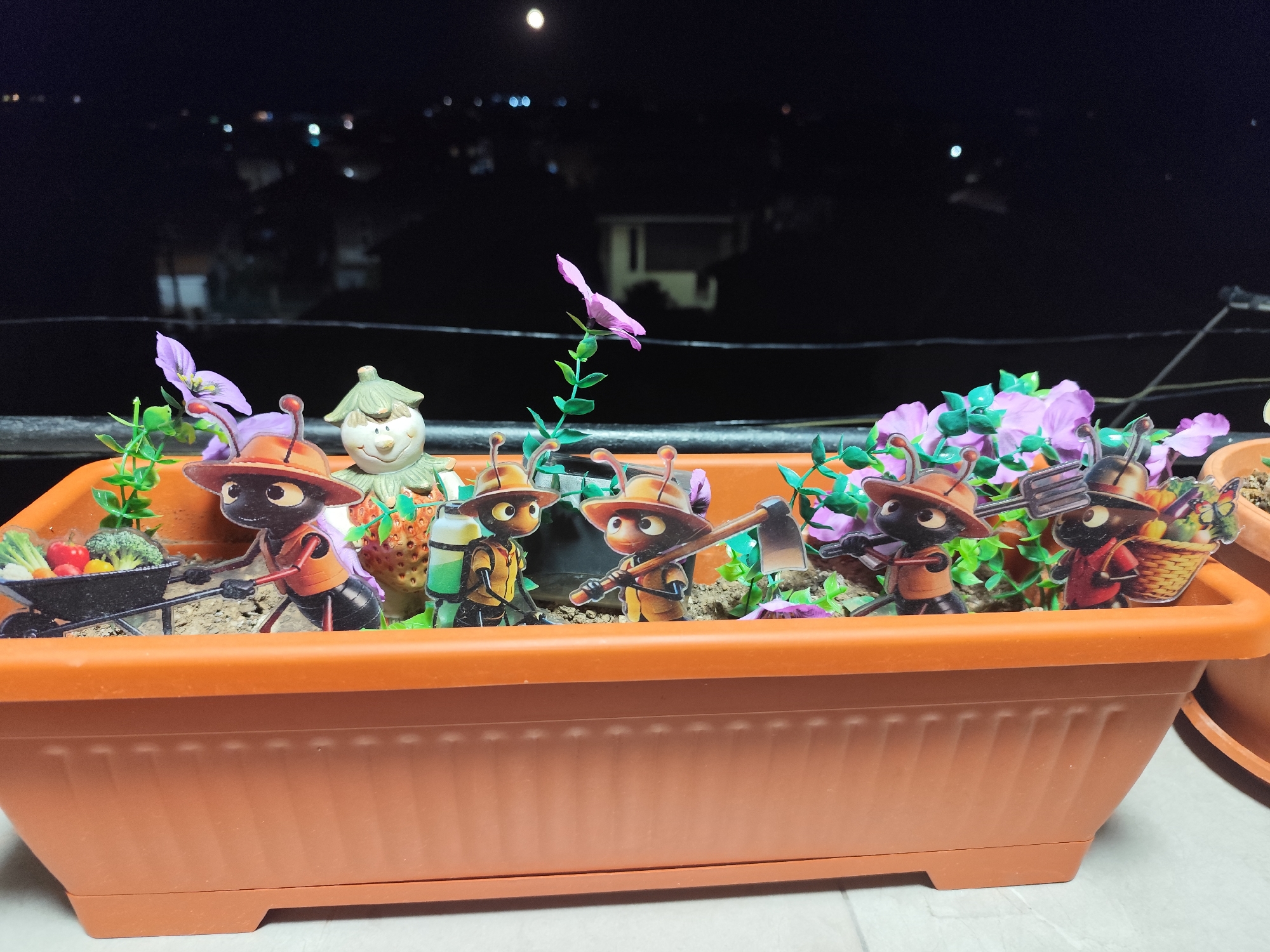In a rectangular orange planter filled with soil and decorative plants, a diorama depicts several cartoon ants dressed in gardening attire. The ants are engaged in various activities, such as pushing a wheelbarrow filled with vegetables, holding gardening tools like a hoe and a pitchfork, and carrying a basket. Behind the ants, a small scarecrow figure stands, and a few artificial purple flowers are scattered throughout the planter. The planter is situated on a railing outdoors at night, with a blurred cityscape visible in the background, including distant lights and what appears to be the moon. The setting suggests a balcony or terrace in a residential area. The overall scene is a whimsical, staged arrangement, not a depiction of real-life events. The city of Montana, Bulgaria is not directly depicted in the image, nor is there any specific information in the visual evidence to place the scene within that location.