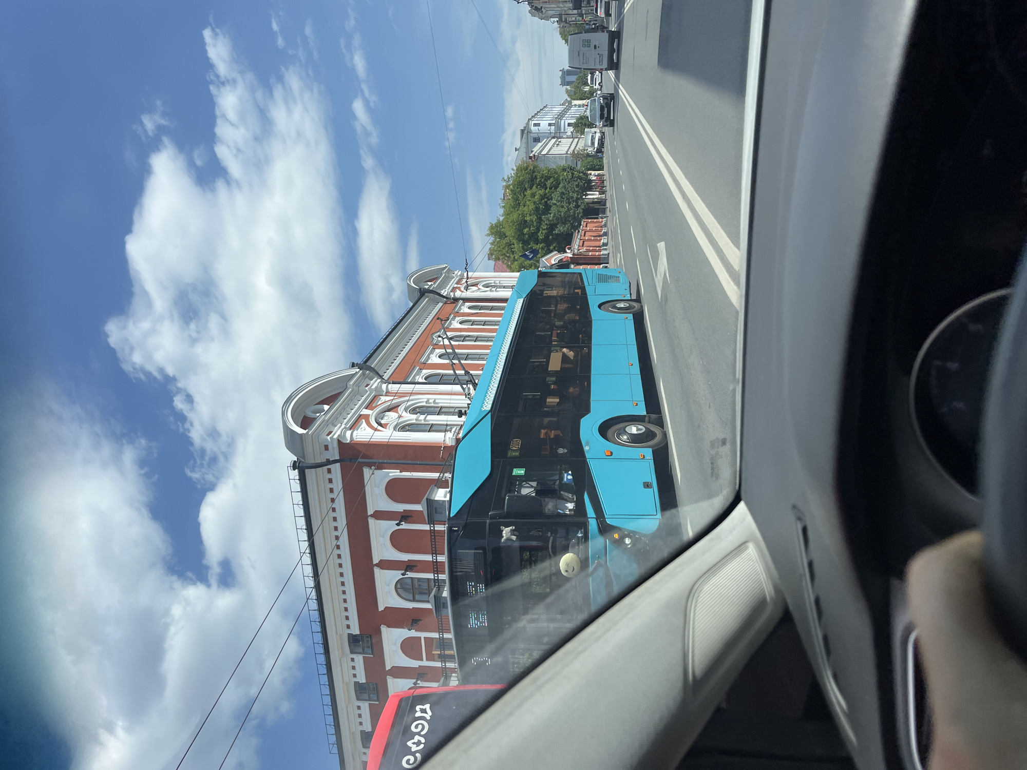 An exterior, eye-level, long shot captures a street scene in Kazan, Russia on a sunny day with scattered clouds. The perspective is from inside a vehicle, looking forward through the windshield, with the dashboard and a person's hand on the steering wheel visible in the foreground. A bright blue trolleybus with black-tinted windows is the dominant subject, driving on the right side of the road. The trolleybus has a white smiley face sticker and a small white plush toy hanging from its interior. Next to it, a red bus with Cyrillic lettering is partially visible.
Behind the buses, a historic building with a red facade, white decorative trim, and arched windows lines the street. Further down the road, other buildings and trees are visible. The road is paved with white lane markings and directional arrows. Several other vehicles, including a white van and a black car, are also on the road. Overhead, power lines for the trolleybus stretch across the sky. The overall impression is a snapshot of urban life in Kazan.
