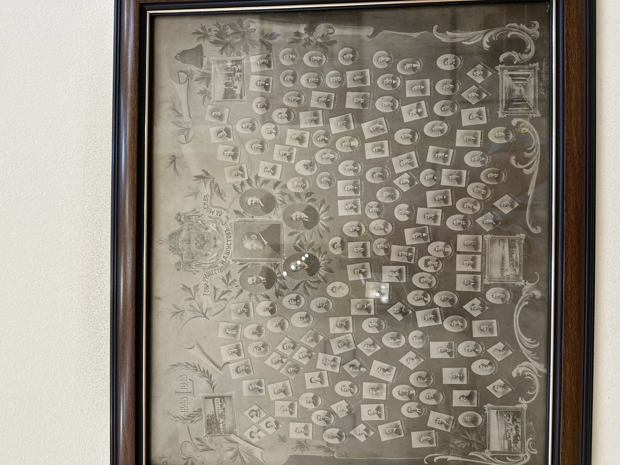 A framed, antique monochrome photographic collage, likely celebrating an anniversary, hangs on a light-colored wall. The dark wooden frame has a black inner trim. The collage itself is intricate, featuring numerous small, circular, oval, and rectangular portraits of men in formal attire, arranged decoratively around larger central portraits and architectural images. The overall tone is faded sepia or monochrome, with some glare and reflections visible on the protective glass.
The central activity captured is a commemoration, evidenced by the dates "1863 1913" displayed prominently at the bottom left, suggesting a 50-year jubilee. Visible Cyrillic text identifies the organization as "ТОВАРИЩЕСТВО Н.Л.ШУСТОВЪ" (Partnership of N.L. Shustov), a prominent Russian company, likely referring to the Shustov Brothers' vodka and cognac distilleries. Another partial text reads "ВЪ МОСКВА" (in Moscow), indicating the company's base. Some individual portraits have names written next to them, such as "Ес. Галкинь" (Es. Galkin).
The scene is a formal portrait collection, possibly representing company founders, employees, or significant associates over its operational period. Decorative elements like leaves, scrolls, and other embellishments frame the various photographic inserts. The architectural images depict buildings or interiors, possibly company premises. The collage reflects a specific historical period, likely early 20th century. The photograph is old and shows signs of age, including fading and possible dust or scratches on the surface beneath the glass.