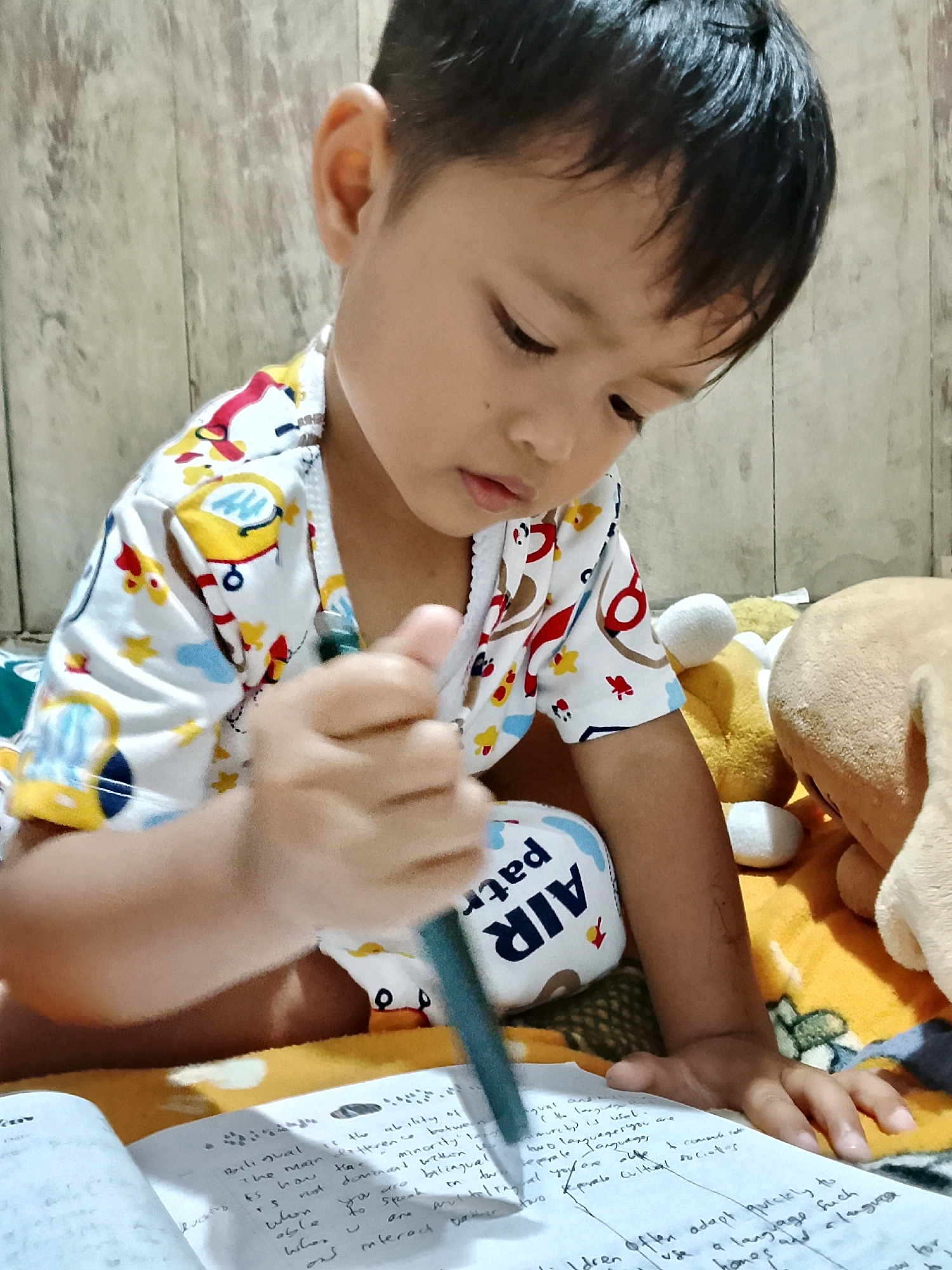 A young boy, likely of Indonesian ethnicity based on the location provided, is shown from the chest up, engrossed in writing in a notebook. He is wearing a white t-shirt with a colorful pattern of cartoon characters. The boy is holding a green pen in his right hand, poised over an open notebook filled with handwritten text. His left arm is resting on a stuffed toy. In the background, there are other stuffed toys and a wall with a wood-like texture. The overall scene appears to be indoors, possibly in a residential setting in Kradenan, Indonesia.
