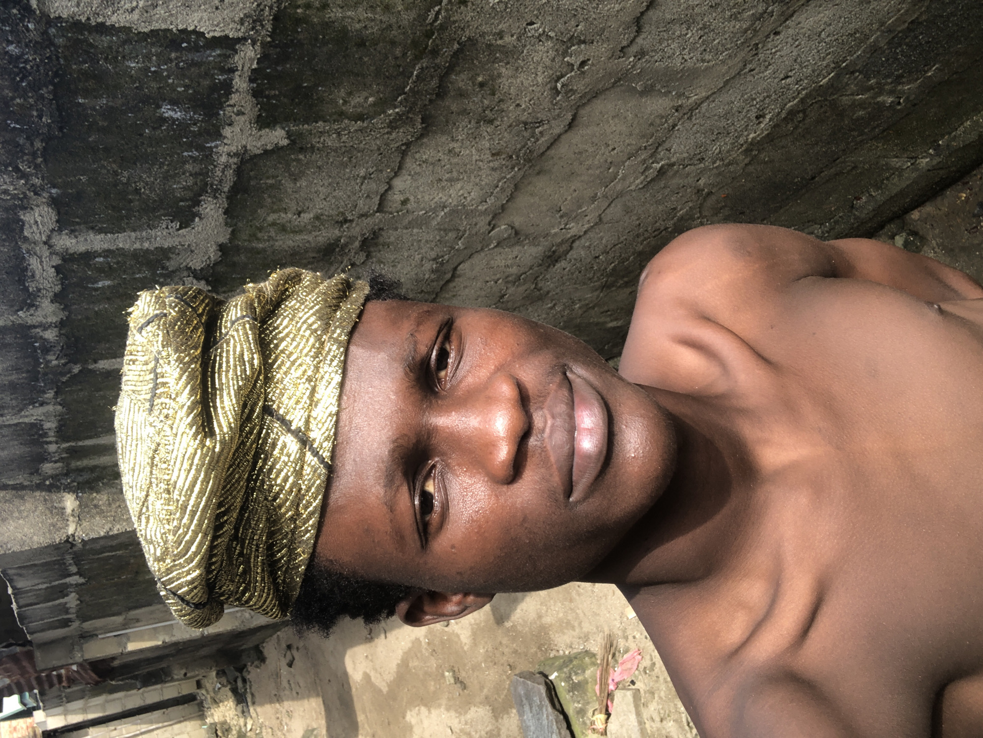 A young Black male, bare-chested, stands in what appears to be a backyard or alleyway in Ado, Nigeria. He is wearing a gold, textured headwrap that covers his hair and sits high on his forehead. His skin is dark brown and appears slightly shiny, possibly from sweat or oil. He has a neutral expression on his face, with his mouth slightly open. His shoulders are visible, showing his collarbones and the curve of his upper arms.
The background consists of a rough, grey concrete wall with patches of green moss or algae growing on it. To the left of the man, there is a dirt path with some scattered debris, including rocks and what looks like some pink material. In the distance, another wall or structure is visible, also made of concrete blocks. The lighting suggests it is daytime, with sunlight casting shadows and highlighting the textures of the wall and the man's skin.