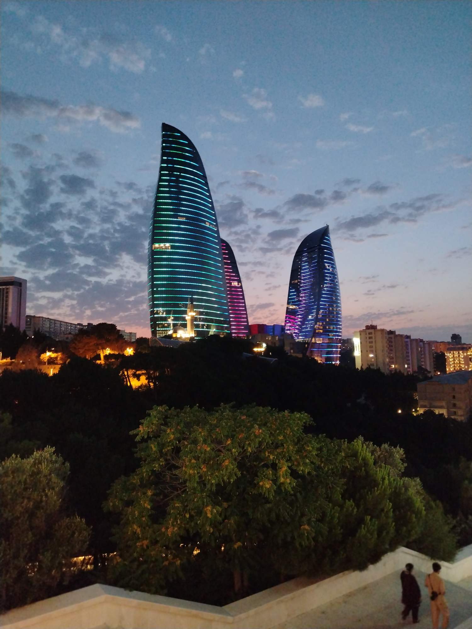 The image captures the Flame Towers in Sabail Raion, Azerbaijan, at dusk. The three modern skyscrapers, shaped like flames, are illuminated with vibrant blue, green, and pink lights that contrast with the darkening sky. The clouds in the sky are wispy and grey, suggesting the transition from day to night. In the foreground, lush green trees obscure the lower portions of the buildings, creating a layered effect. A white stone staircase leads downwards, and two individuals, one in dark clothing and the other in light-colored attire, are walking down the steps. The background reveals a city skyline with other buildings, some also illuminated. The overall scene is a blend of modern architecture and natural elements, bathed in the soft light of evening.