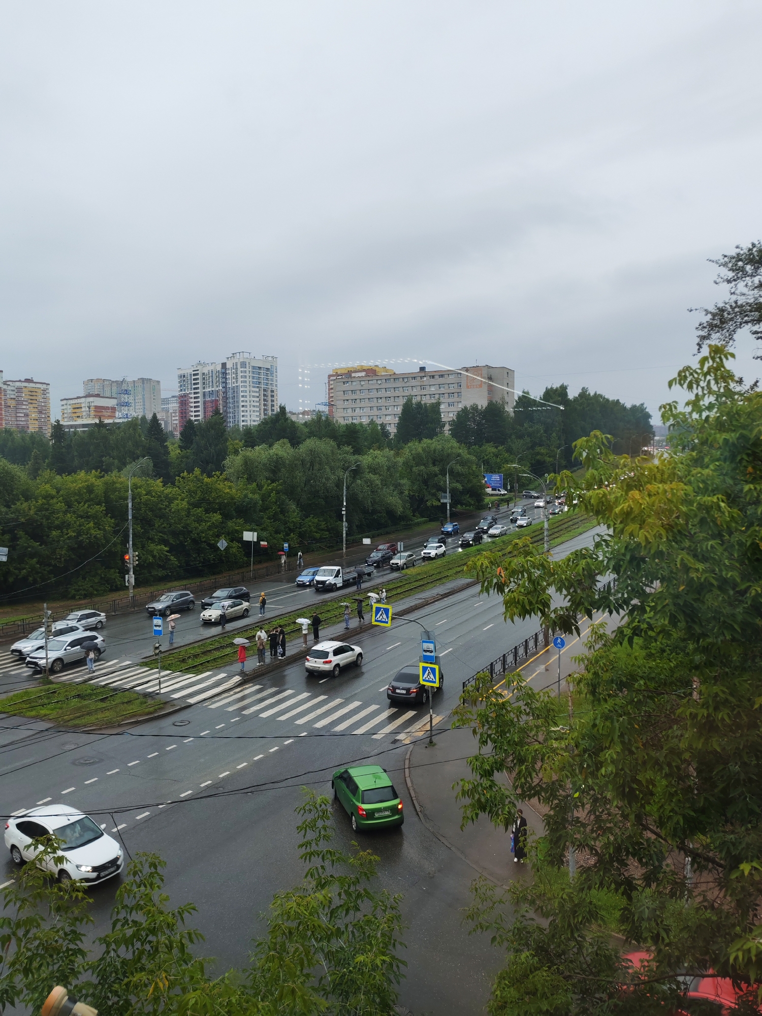 The image depicts a street scene in Izhevsk, Russia, on an overcast day. A multi-lane road runs through the frame, wet from recent rain, with several cars, including a white hatchback, a green station wagon, and other vehicles in traffic. Pedestrians, some holding umbrellas, are visible on the sidewalks and at crosswalks. Tall residential buildings and lush green trees line the street. Traffic lights and street signs are present. The overall atmosphere is subdued, typical of cloudy weather.