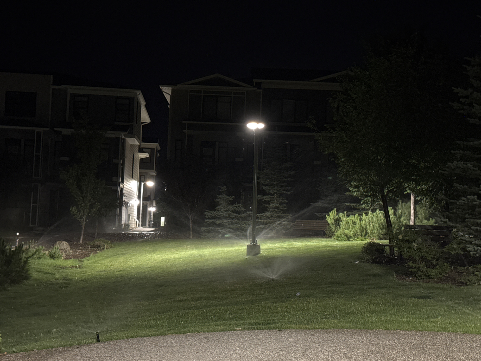 This is a night-time photograph of an irrigated lawn in front of a multi-story apartment complex. The buildings are modern in style with dark facades and illuminated windows. A streetlamp illuminates the central area of the lawn, where sprinklers are actively watering the grass, creating a misty effect. Several trees and bushes are present on the property, as well as a wooden bench visible in the background. The foreground shows a gravel pathway. The scene is located in a residential area of Calgary, Canada, specifically in the community of University Heights.