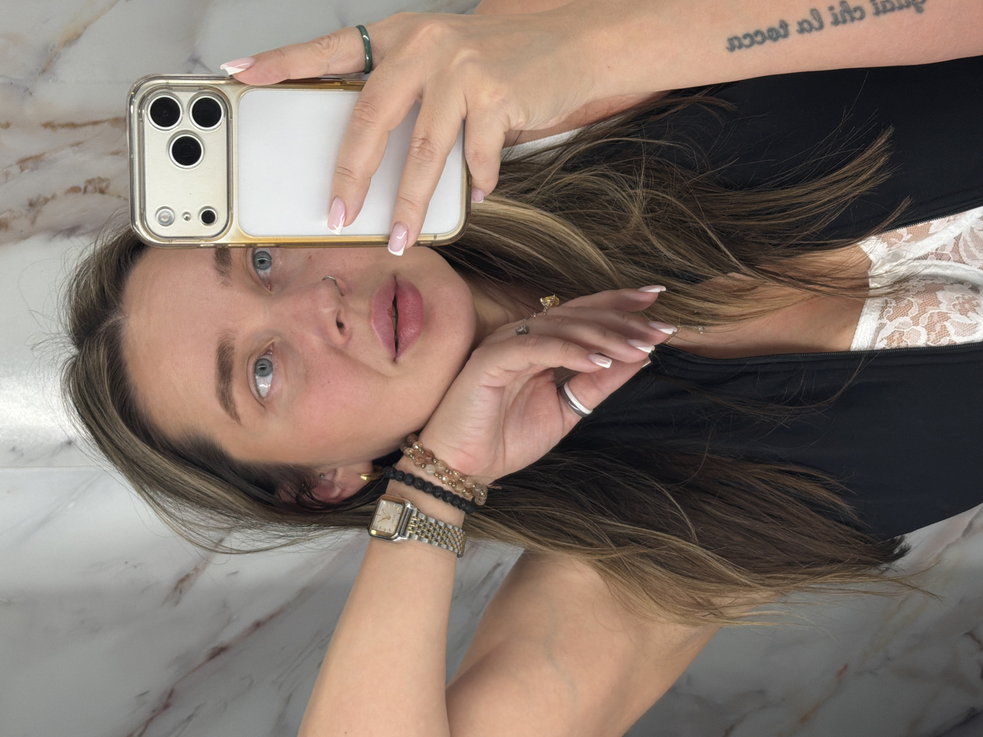 A person is taking a selfie in front of a marble background. The person has fair skin, light brown hair with blonde highlights, and blue eyes. They are wearing a black top with a white lace undershirt. The person is holding a smartphone with a clear gold-trimmed case and a white back over their forehead. They are wearing a green ring, a gold ring, a silver ring, a watch, and beaded bracelets. The person has a nose piercing and a tattoo on their arm. The location is not identifiable as Podolsk, Russia, based on the image.