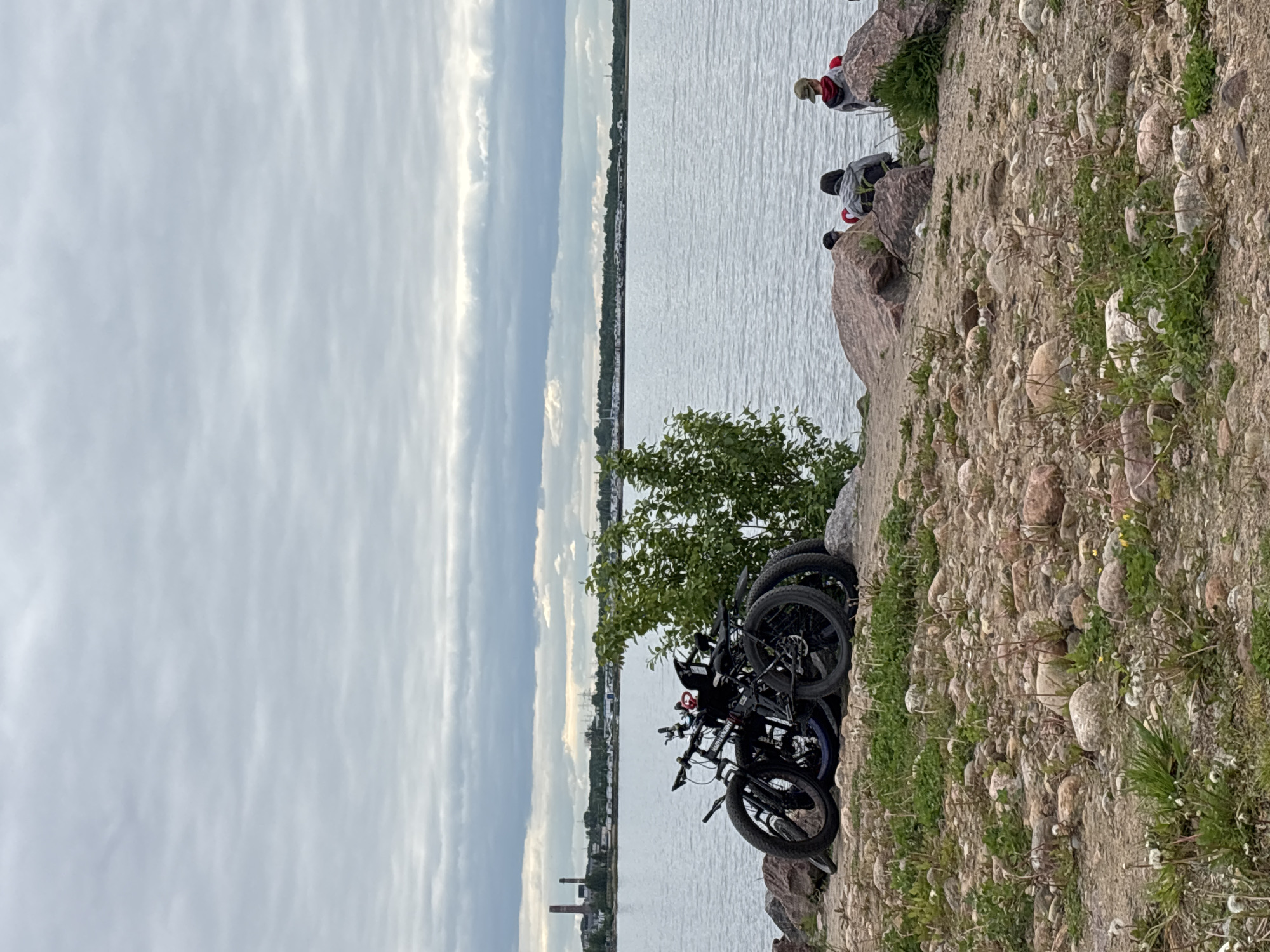 The image depicts a waterfront scene, likely in Saint Petersburg, Russia, given the distinctive cityscape and the geological features of the landscape. The foreground features three dark-colored bicycles parked on a rocky, uneven terrain interspersed with small patches of green vegetation. The rocky area leads down to a body of water, likely the Gulf of Finland or a portion of the Neva River. In the middle ground, across the water, is a built-up area, possibly Kronstadt, characterized by urban structures including a smokestack and buildings. The sky is overcast, with layers of gray and white clouds. At the upper right corner, there are a few individuals seated on the rocks, overlooking the water.