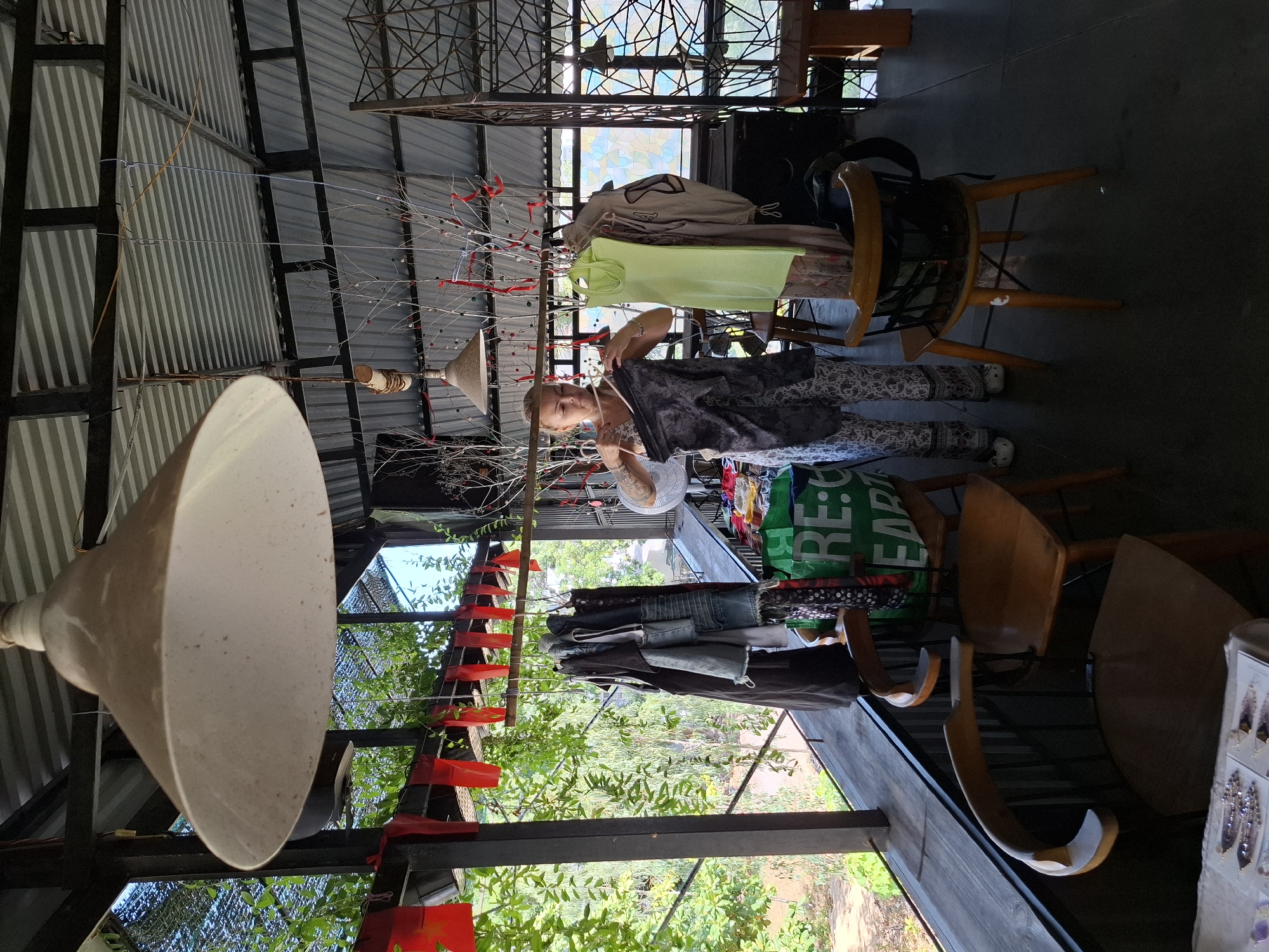 A woman stands indoors, examining a dark garment she holds. She is wearing patterned pants and white shoes. The setting appears to be an open-air structure with a corrugated metal roof and open sides. To the left, a rack of dark clothing hangs, and beyond it, lush green foliage is visible through a railing. To the right, several chairs are placed around tables, with some clothing items and a green bag with text that reads "RE: EARTH" visible. The overall scene suggests a market or a retail space with an outdoor ambiance. This location is likely a roadside stall or a small shop in the coastal city of Nha Trang, Vietnam, characterized by its tropical vegetation and semi-open structures designed to take advantage of the climate.