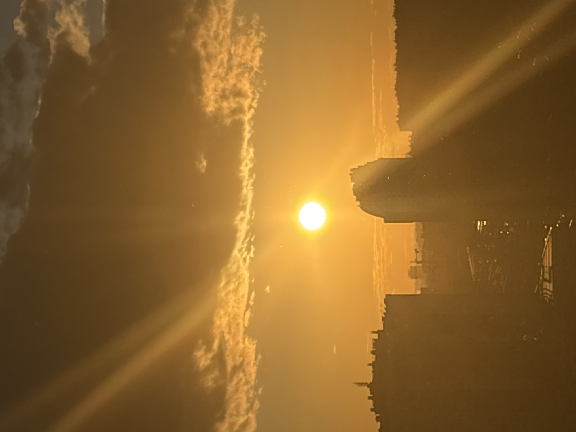 The image captures a sunset over a cityscape, likely in an urban environment. The sun, a bright, glowing orb, is positioned slightly to the right of the center in the frame, casting a warm, golden light across the scene. Sunbeams radiate from the sun, cutting diagonally through the frame, creating a dramatic effect. Wispy clouds, tinged with orange and yellow hues from the setting sun, are scattered across the sky, adding texture and depth.
In the foreground, the silhouettes of buildings are visible against the bright sky. On the left, a row of multi-story buildings, appearing to be apartment blocks or office buildings, forms a dark silhouette. A prominent building with a curved roof and distinct architectural features stands out in the center, also in silhouette. Further in the background, more buildings and a treeline are faintly discernible through the haze and the golden light. The overall impression is one of a tranquil, albeit somewhat hazy, urban sunset. The city's infrastructure, such as streets or railway tracks, can be vaguely made out in the lower portion of the image, softened by the atmospheric conditions.