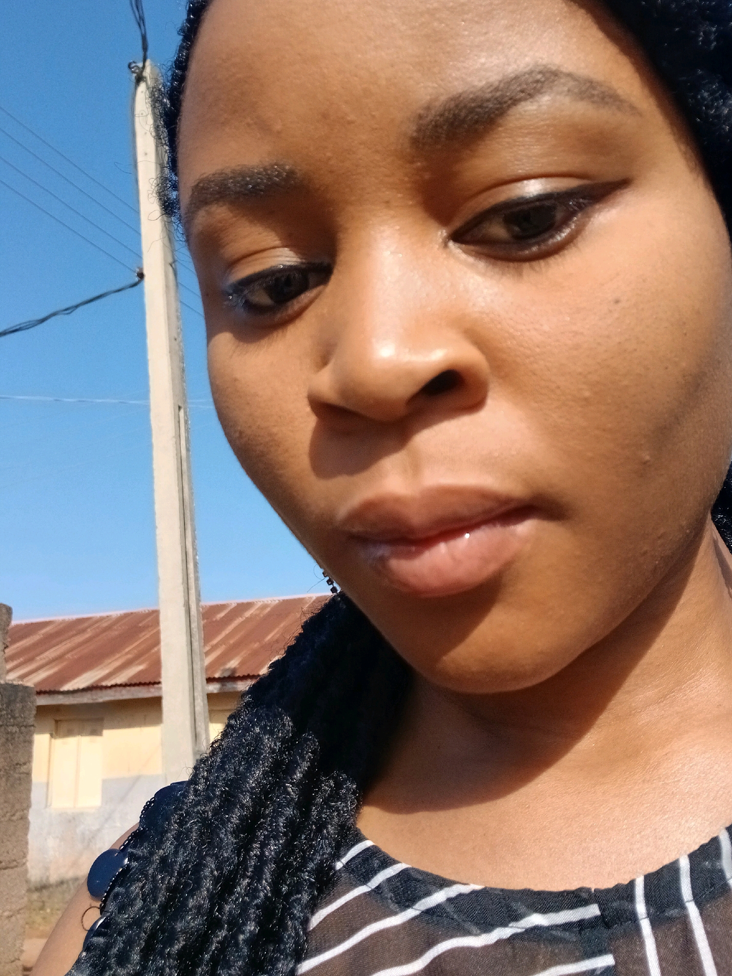 A medium close-up, slightly downward angled selfie shows a young Black woman outdoors. She is facing the camera, with her face filling most of the frame. Her skin is a warm brown tone, and she has dark, braided hair that falls over her shoulder. She is wearing a black and white striped top. Her left eye is visible, and she has on dark eyeliner and what appears to be mascara. Her lips are slightly parted and have a subtle sheen. Behind her, a clear blue sky is visible with a utility pole and some power lines running diagonally across it. In the background, the roof of a building with corrugated metal sheeting is visible, along with a section of a yellow wall. The scene appears to be in a residential area, possibly Gura Top, Nigeria, given the architectural style and general environment. The lighting suggests it is daytime, with harsh sunlight casting shadows.
