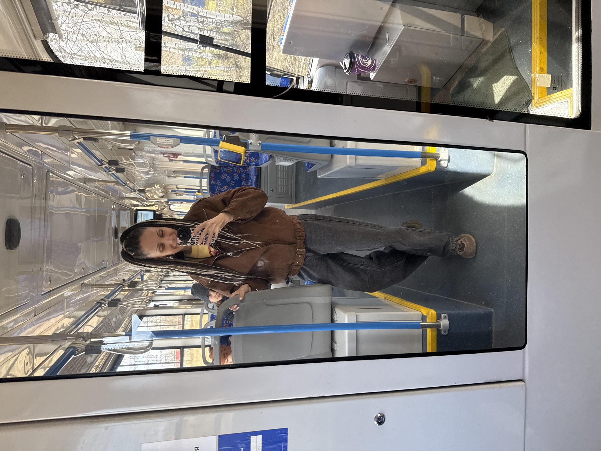 The image captures a mirror selfie taken by a person inside a public transport vehicle, likely a bus or tram, during daylight hours.
The primary subject is a person standing, reflected in a large window. They have long, dark dreadlocks and are wearing a brown jacket, a yellow top, dark jeans, and brown boots. They hold a smartphone to capture the reflection, smiling subtly. The phone case has a distinct black and white pattern.
The interior of the vehicle is visible, featuring numerous silver handrails and blue accent bars, along with hanging white straps for standing passengers. Blue patterned seats are arranged along the aisle. The floor is dark with bright yellow safety strips. A purple and white swirled cup rests on a surface near a window. A small grey cloth is tucked into the upper corner of a window frame in the top right. Reflections of other passengers, including a child in a beanie, are partially visible in the windows and interior surfaces.
Outside the windows, the view consists of bare tree branches against a bright sky, suggesting a sunny day in late winter or early spring. The strong sunlight casts bright reflections and highlights within the vehicle.
The activity is the person taking a full-body mirror selfie. The setting is a public transport ride in Rusanovo, Russia, as indicated by the context.
Visible text includes a pattern/text on the person's phone case, and partially visible Cyrillic text on a blue sticker affixed to the lower part of the bus interior, notably beginning with the letter "Я".