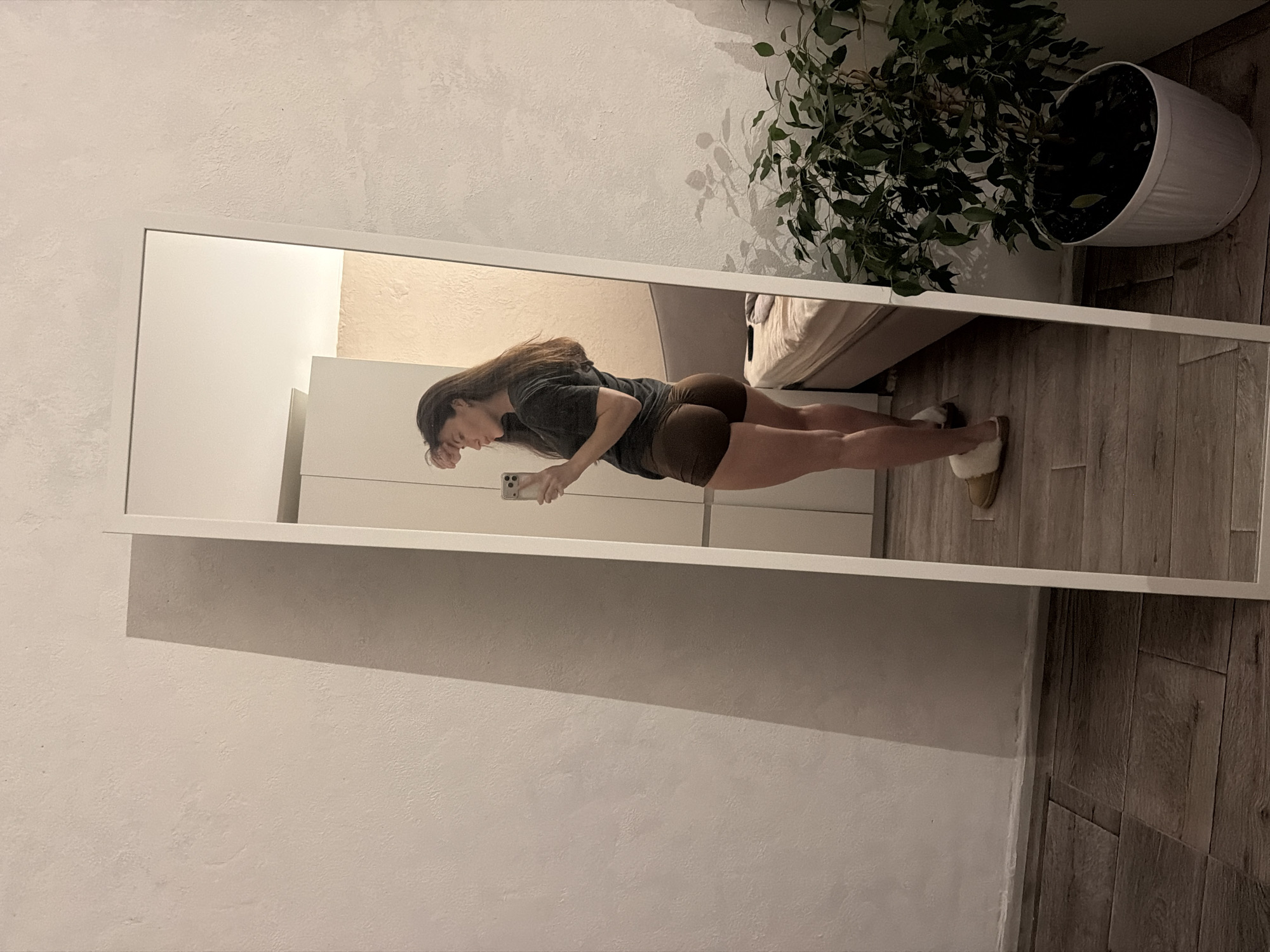 A woman is standing in a room, reflected in a full-length mirror with a white frame. She is holding a phone in her hand. The room has beige-colored walls and brown flooring. A potted plant is located near the mirror. The location is not discernible from the image.