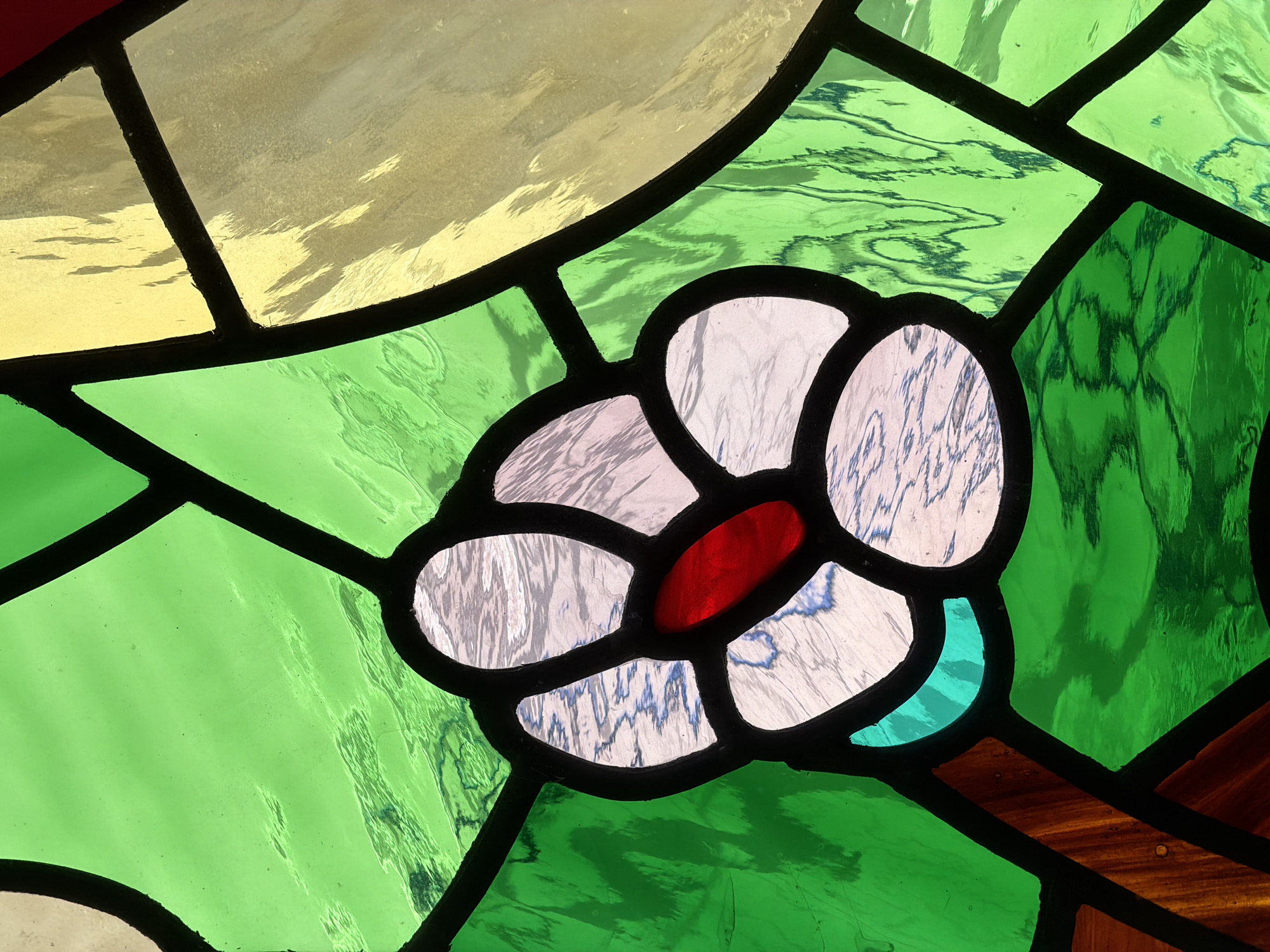This is a close-up, detailed photograph of a stained-glass window, likely located in Arcahueja, Spain. The window features a central floral motif rendered in a variety of translucent colored glass pieces, primarily white with subtle blue veining, a vibrant red center, and surrounded by iridescent white glass petals. These are bordered by thick, dark lead lines. The flower is set against a backdrop of large, irregular panes of green glass, with some panes exhibiting a textured, wavy pattern. In the upper right portion of the frame, sections of pale yellow and creamy colored glass are visible, also bordered by lead. The overall impression is one of intricate craftsmanship and the interplay of light and color characteristic of stained glass artistry, suggesting it is an element of a church, chapel, or historic building in Arcahueja. No people or specific actions are depicted.
